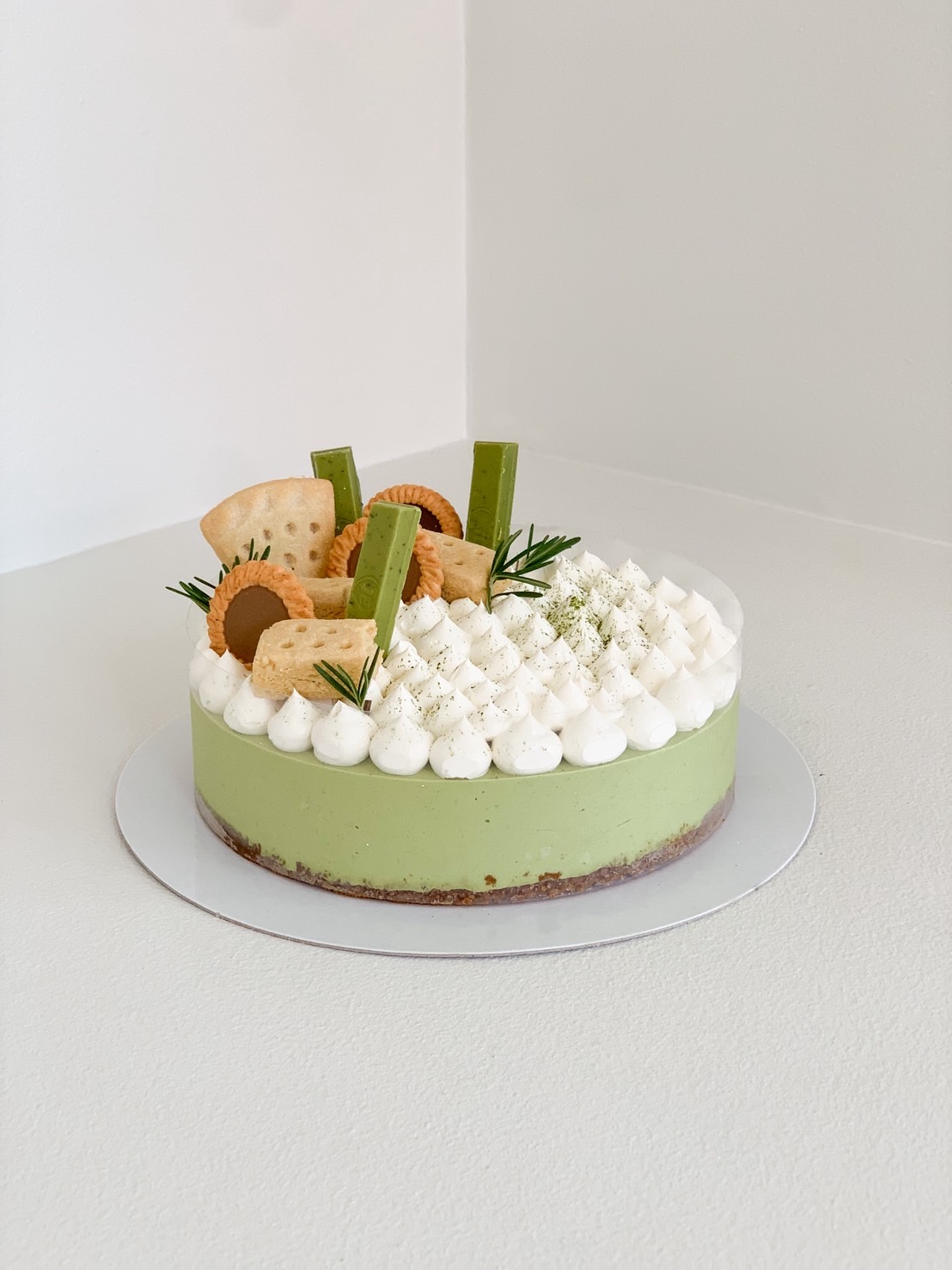 Green matcha mousse cake topped with whipped cream, cookies, rosemary, and matcha chocolate.