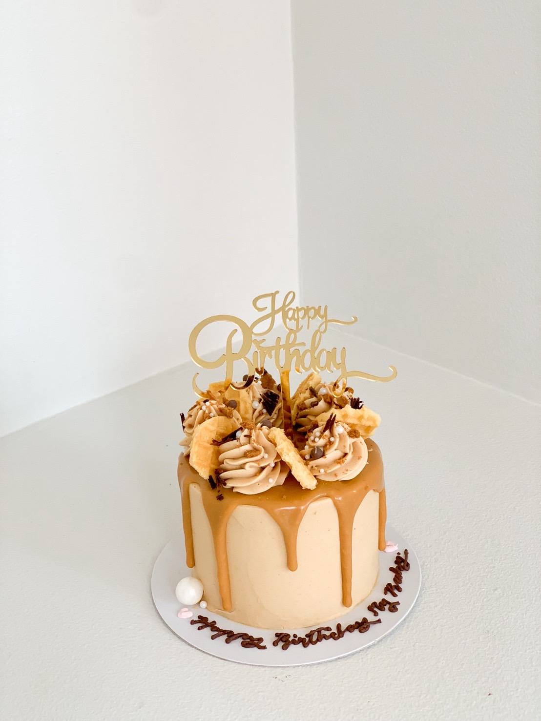 Round caramel drip cake topped with whipped cream, crisps, gold topper, and chocolate crumbs, with birthday message on the base.