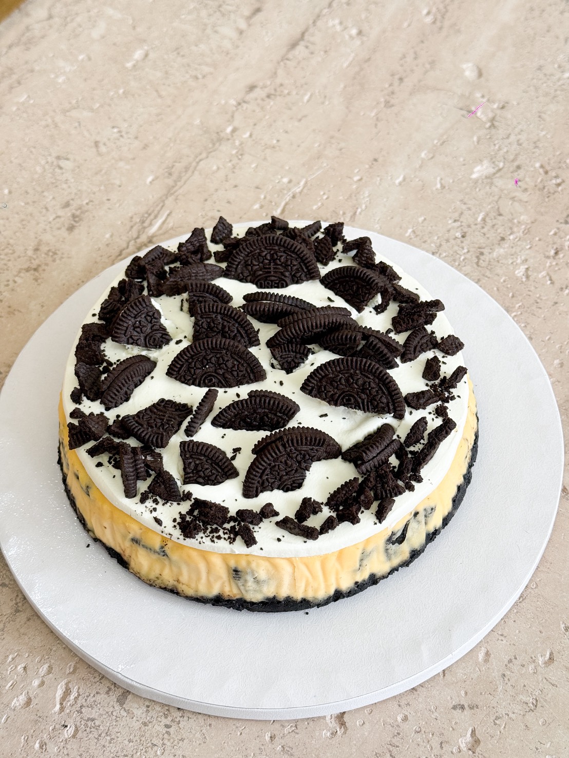 An Oreo Cheese Cream Cake with a dark biscuit base, topped with white cream and scattered pieces of chocolate sandwich biscuits, sits on a white cake board on a light marble surface.