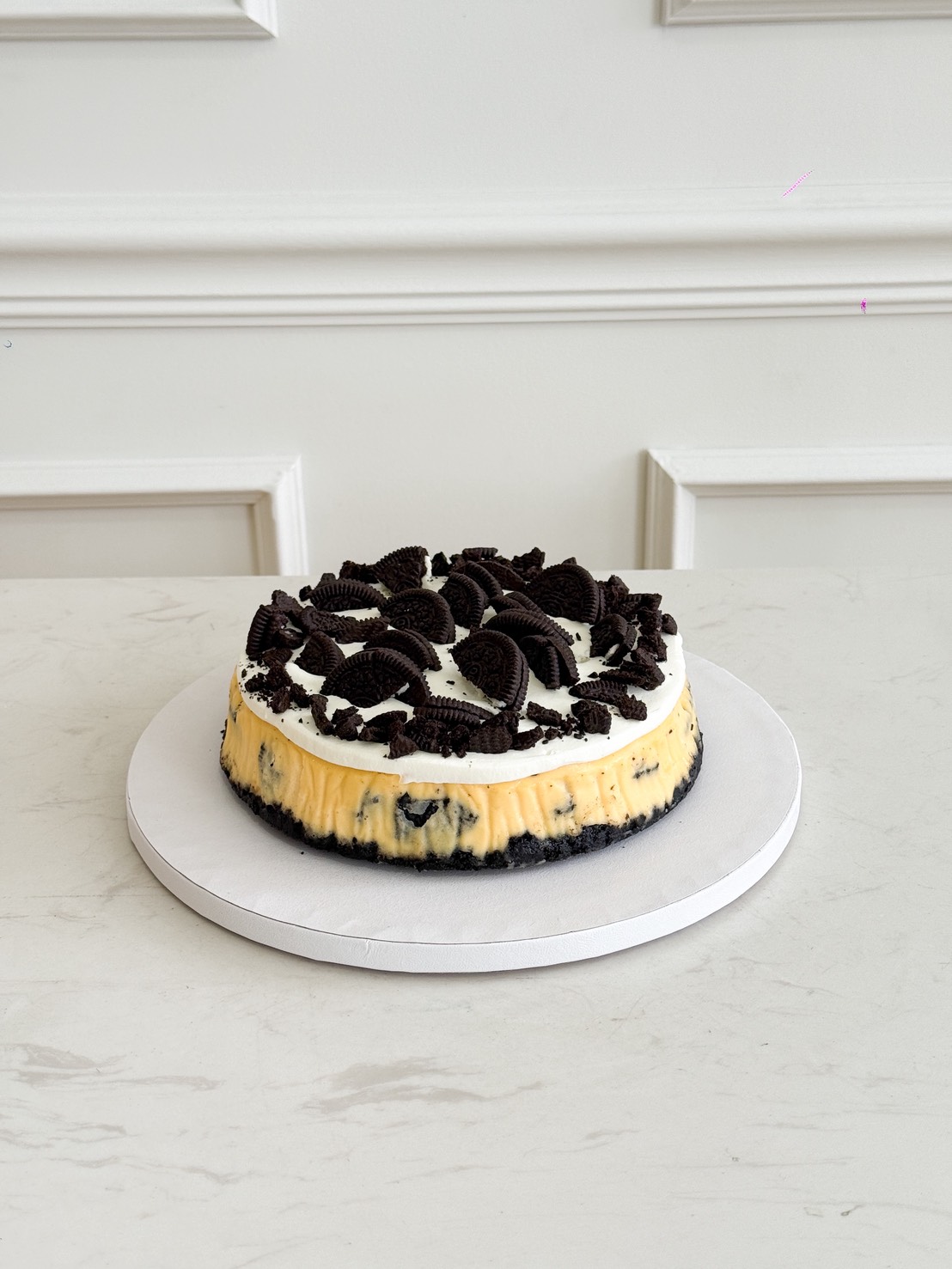 A round Oreo Cheese Cream Cake topped with crumbled chocolate biscuit sandwiches sits on a white cake board, displayed on a light marble surface against a white panelled wall.