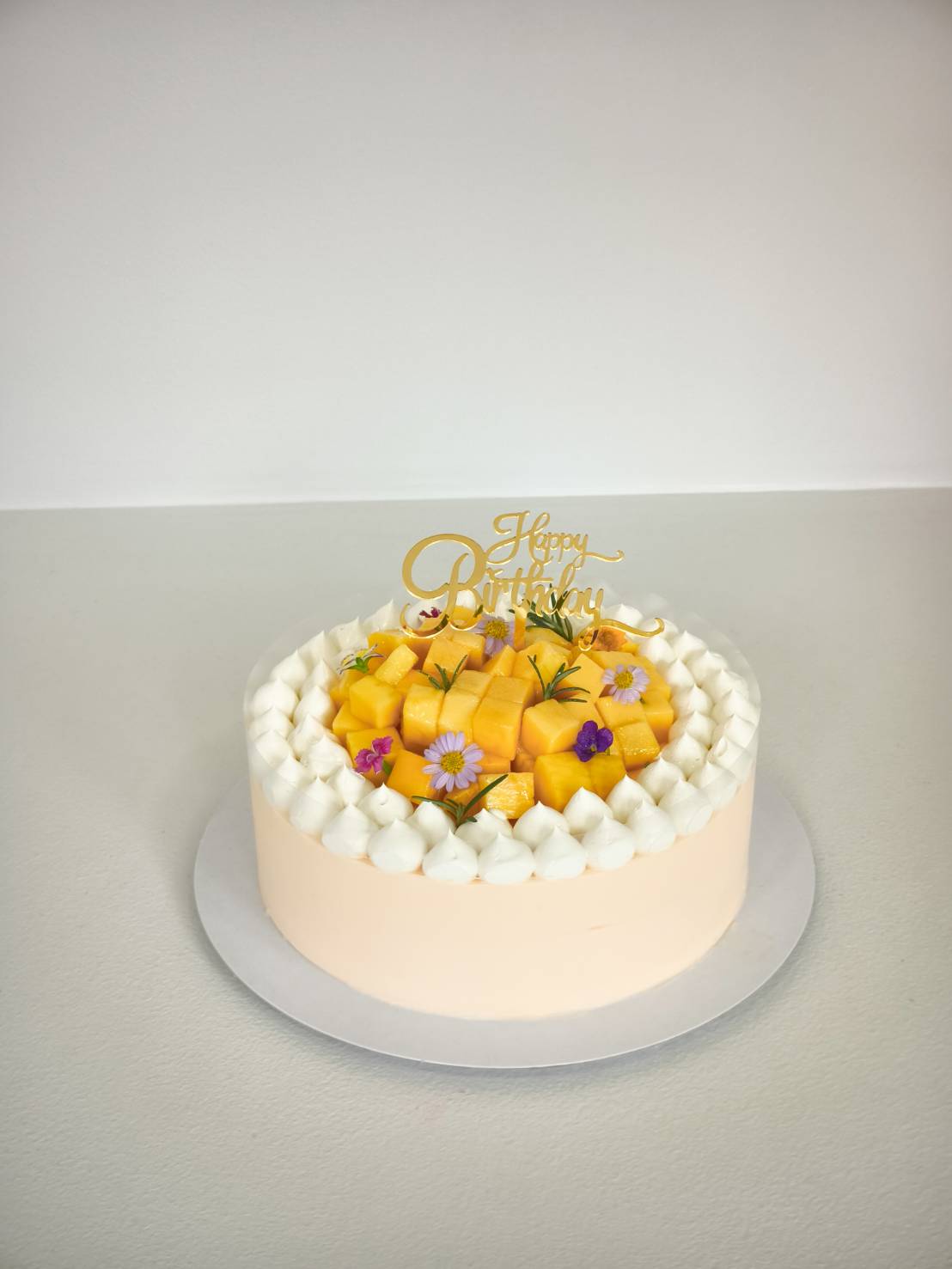 Vanilla cake topped with cubes of fresh mango, whipped cream, and edible flowers, with a Happy Birthday topper.