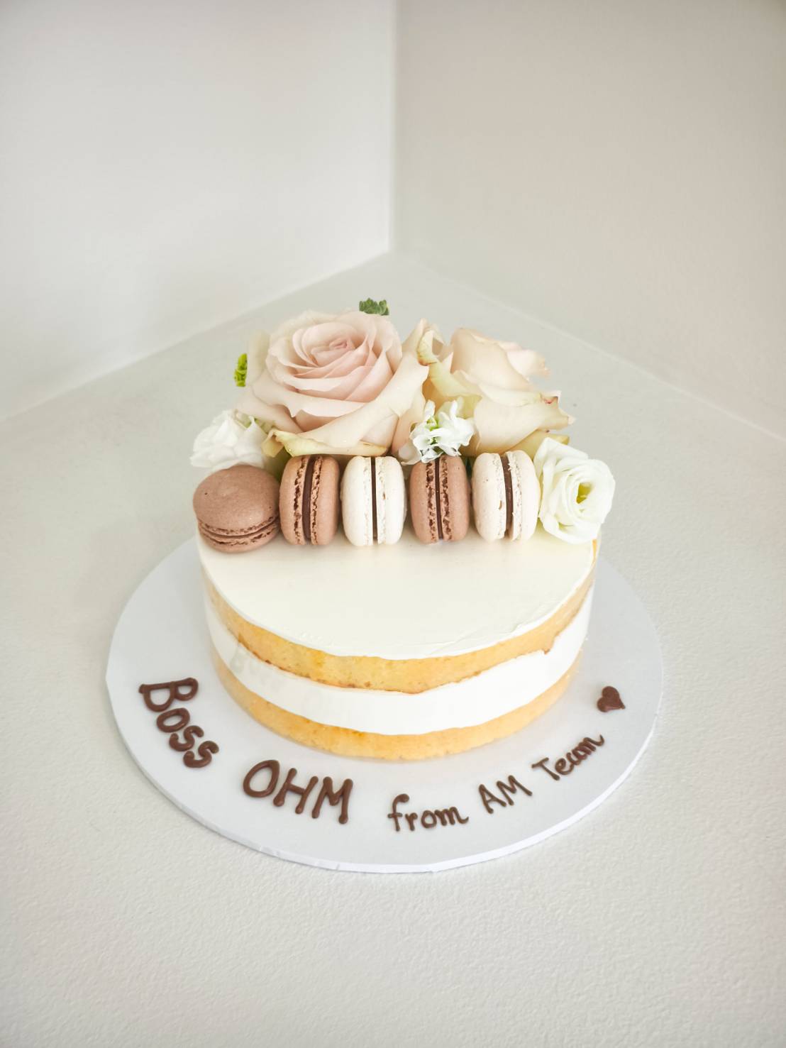 A classic white-iced cake adorned with pink roses, white blooms, and macarons, inscribed for “Boss OHM”—a Charlotte Bakery delight.
