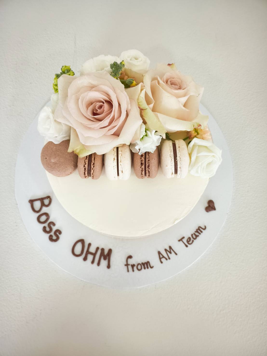 Charlotte Bakery’s signature white frosted cake, crowned with blush roses and macarons, features “Boss OHM from AM Team” in chocolate.