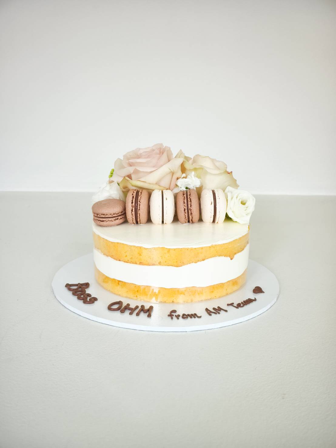 A semi-naked Charlotte cake crowned with fresh roses and macarons, with a heartfelt “DESS OHM from AH Team ♥” in rich icing.
