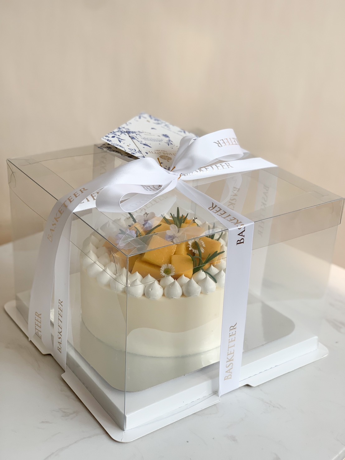 Mango cream cake decorated with whipped cream, fresh flowers, and herbs, presented in a clear gift box with white Basketeer ribbon.