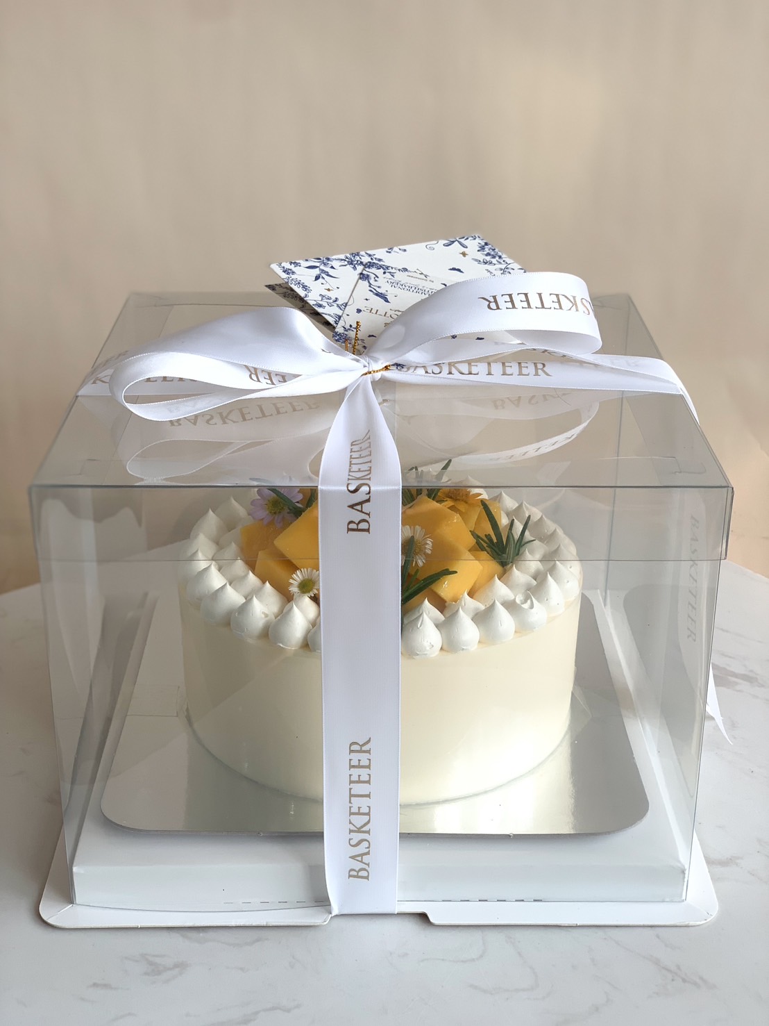 Mango cream cake decorated with whipped cream, fresh flowers, and herbs, presented in a clear gift box with white Basketeer ribbon.