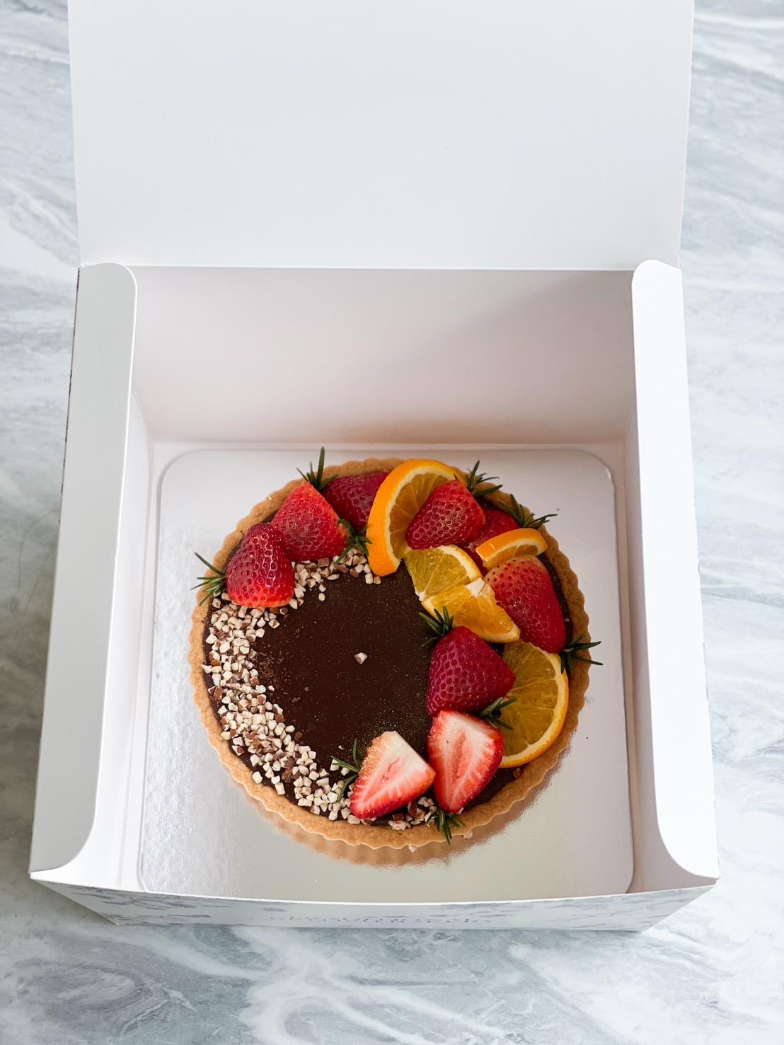 A healthy dark chocolate tart topped with almond bits, fresh strawberries, and orange slices. The fruit tart is placed inside an open white box on a marbled surface.