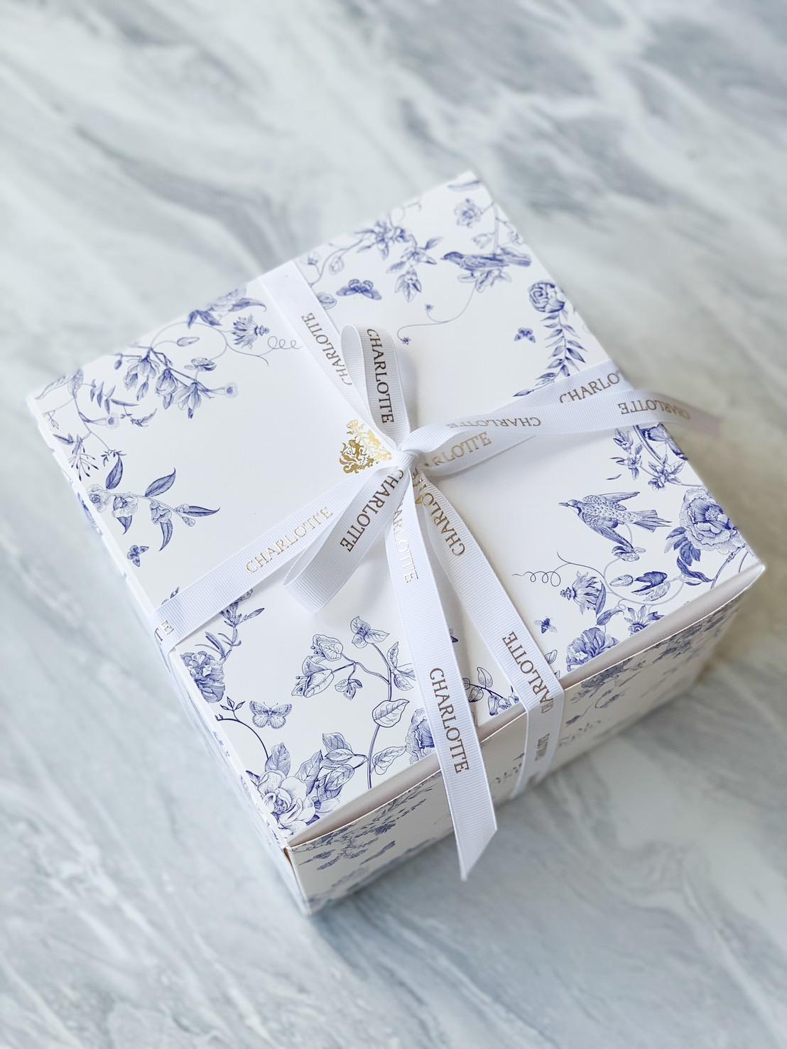 A refined square gift box with blue florals and a “Charlotte” ribbon rests on marbled grey, perfect for elegant Bangkok gifting.