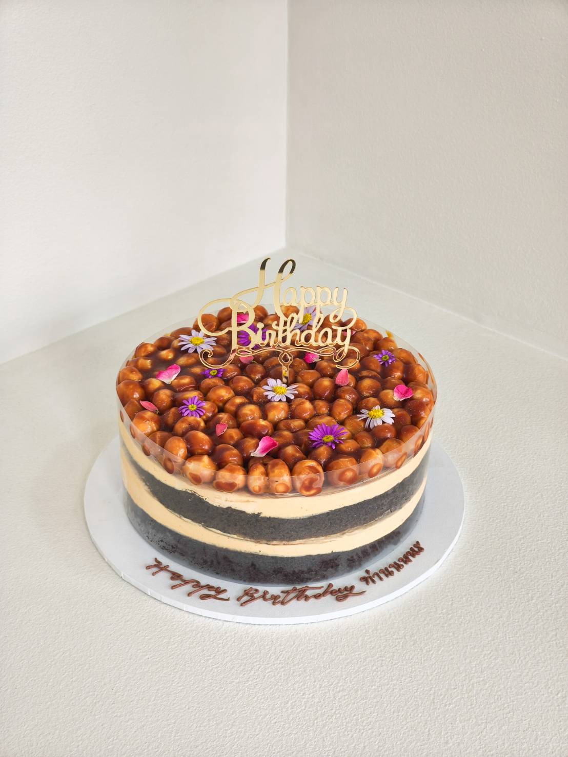 Chocolate caramel layer cake topped with macadamia nuts and edible flowers, decorated with a golden “Happy Birthday” topper.