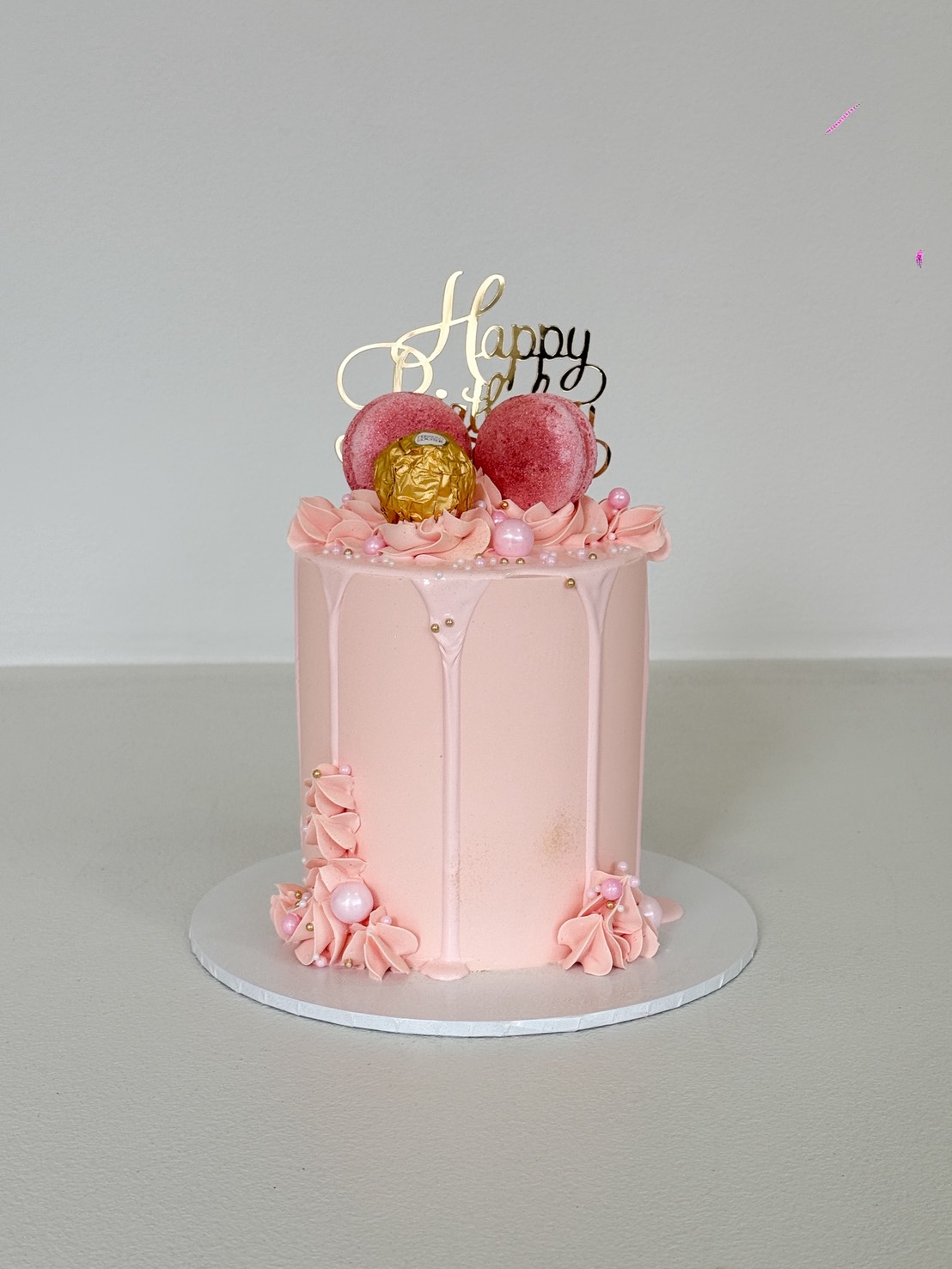 A Blush Drip Delight Layer Cake: a small pink cake with white icing drips, adorned with gold and pink balls, pink flowers, and a gold 