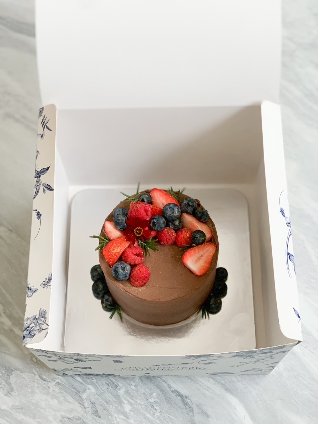 An exquisite Mini Three-Colored Chocolate Cake, adorned with velvety smooth frosting, is elegantly garnished with fresh strawberries, luscious blueberries, and aromatic sprigs of rosemary. This decadent delight rests atop a radiant golden cake board. Nearby, a scatter of fresh fruits adds to the scene's luxurious allure. The distinctive 