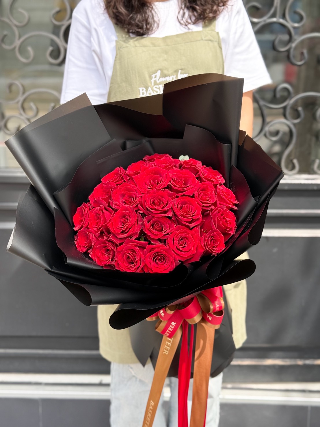 A person wearing a light green apron holds the Eternal Love Red Rose Bouquet, vibrant red roses wrapped in black paper and tied with decorative ribbons, standing in front of a black and white ornate backcloth.