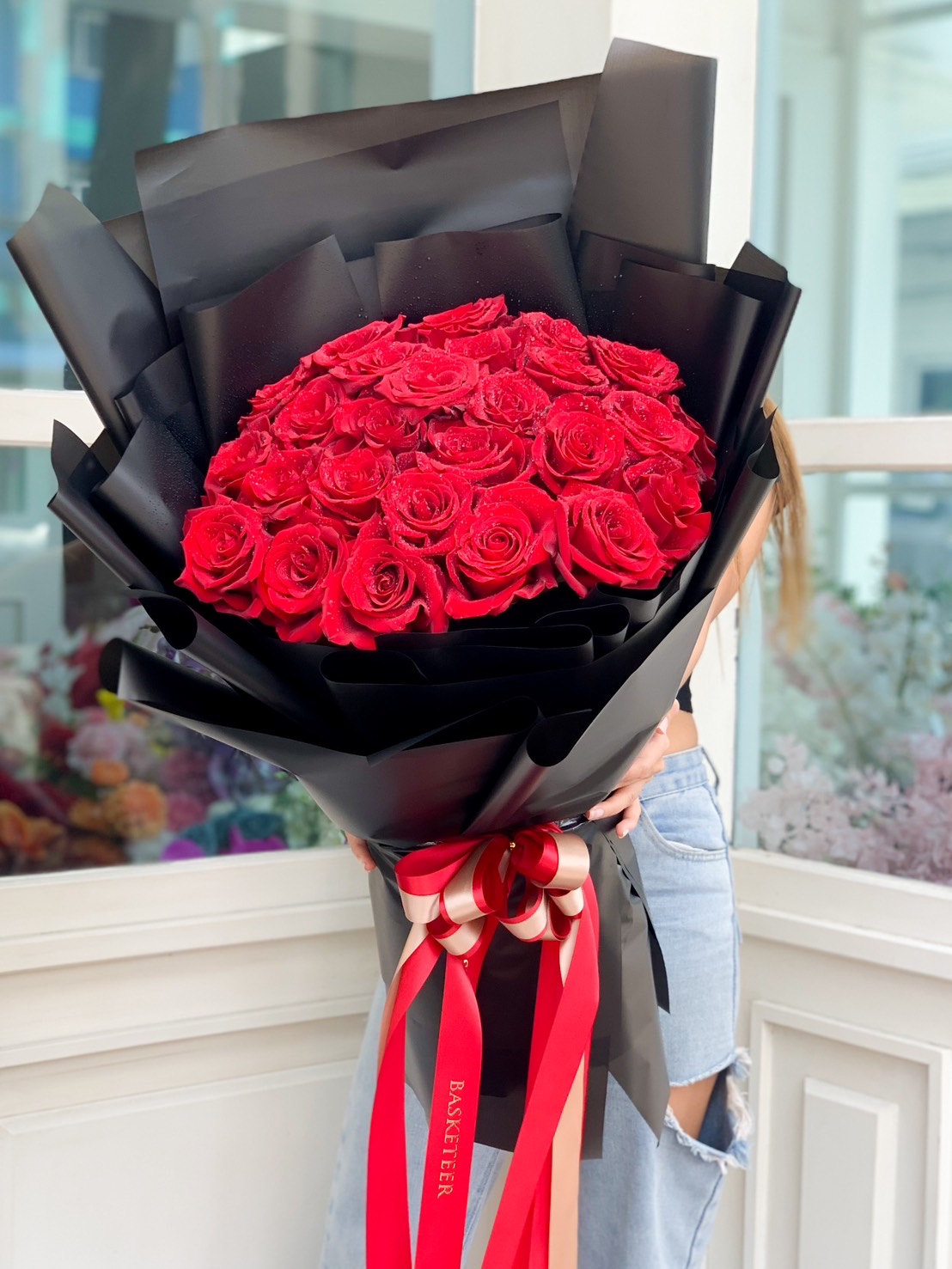 A Love Blossom Explorer is partially obscured by a large bouquet of vibrant red roses wrapped in black paper and adorned with red and beige ribbons. The flowers are arranged in a rounded, densely packed design, creating a stunning and lavish display.