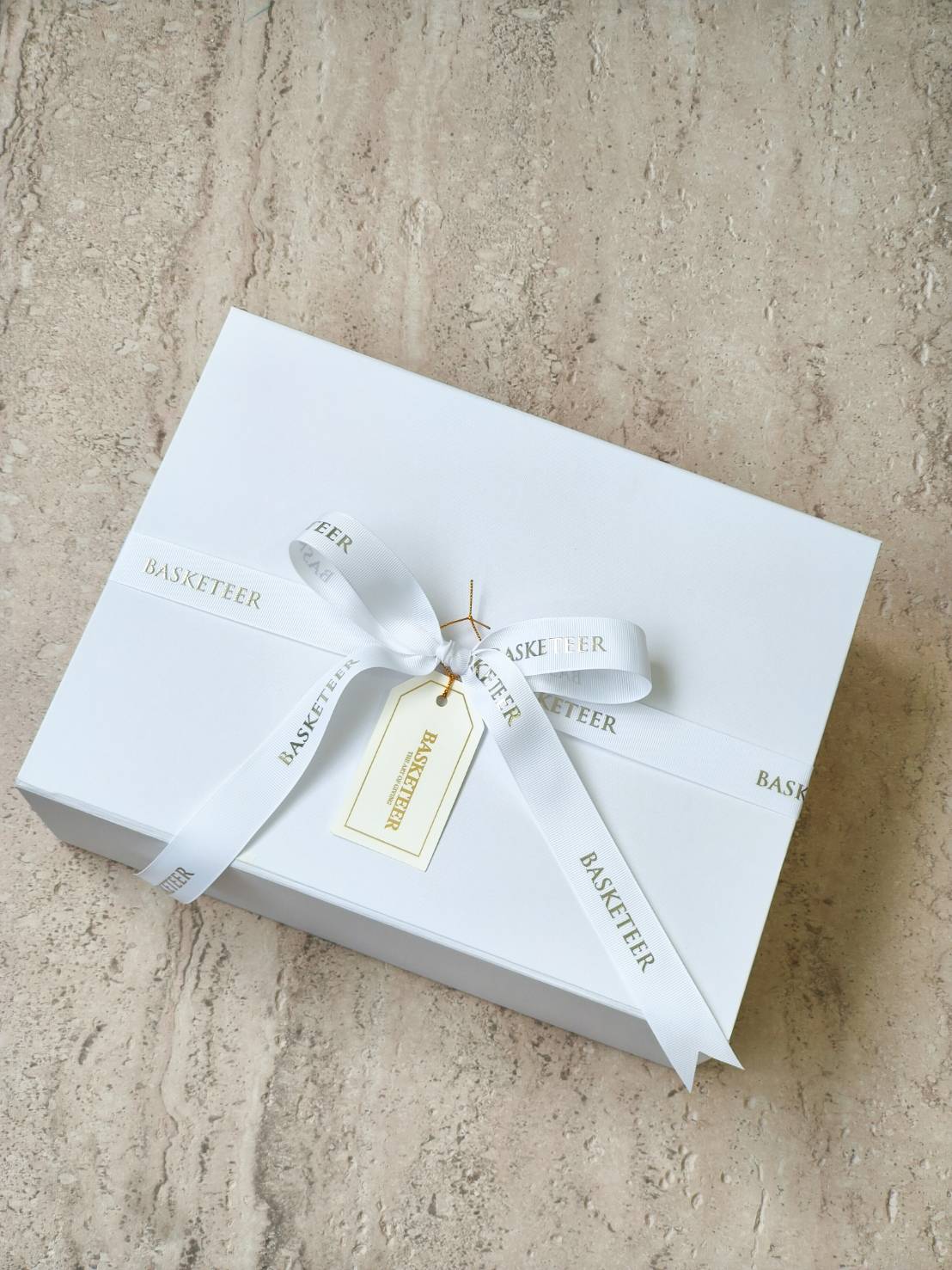 Elegant white gift box tied with white “Basketeer” ribbon and gold-edged gift tag, luxury packaging for premium gifts.