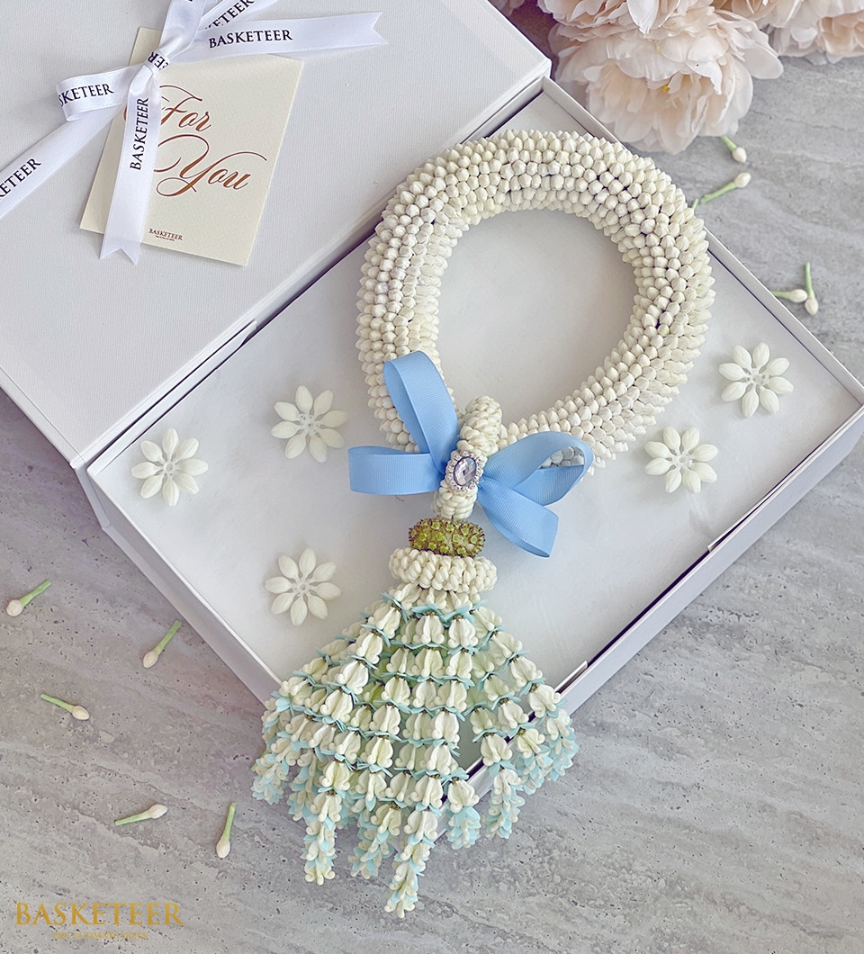 A white floral garland with light blue ribbon and cascading flowers, elegantly arranged inside a white box adorned with delicate flower embellishments. The box is open, revealing a 