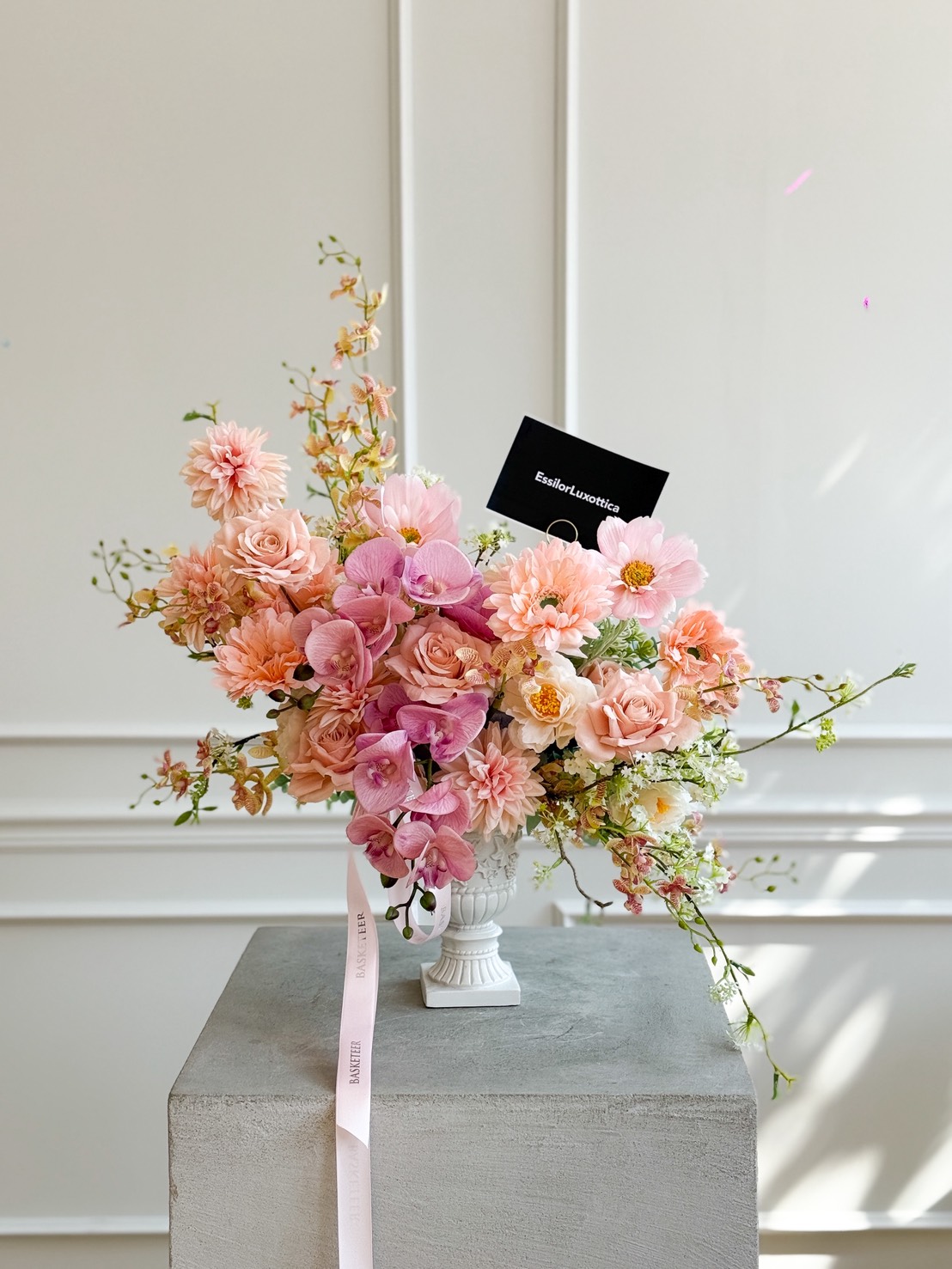 A white vase holds a lush arrangement of artificial flowers in peach tones—roses, dahlias, and orchids with greenery. A black card is nestled among the blooms. The vase sits on a grey pedestal against a white wall.