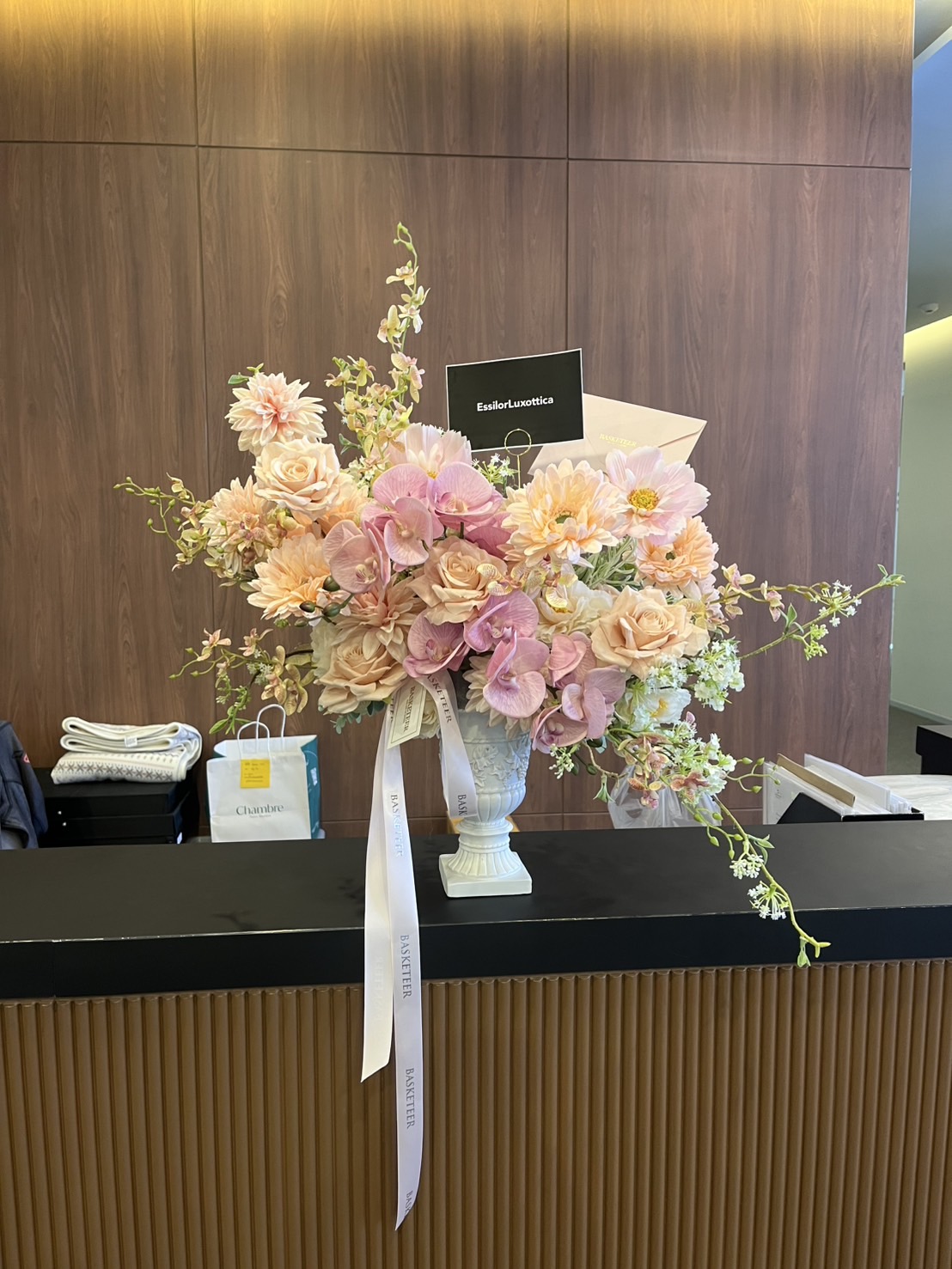 A large bouquet of pastel artificial flowers in vase, featuring pink roses, dahlias, and peach tones, sits on a reception counter with a white ribbon and small sign. A shopping bag is visible in the background.