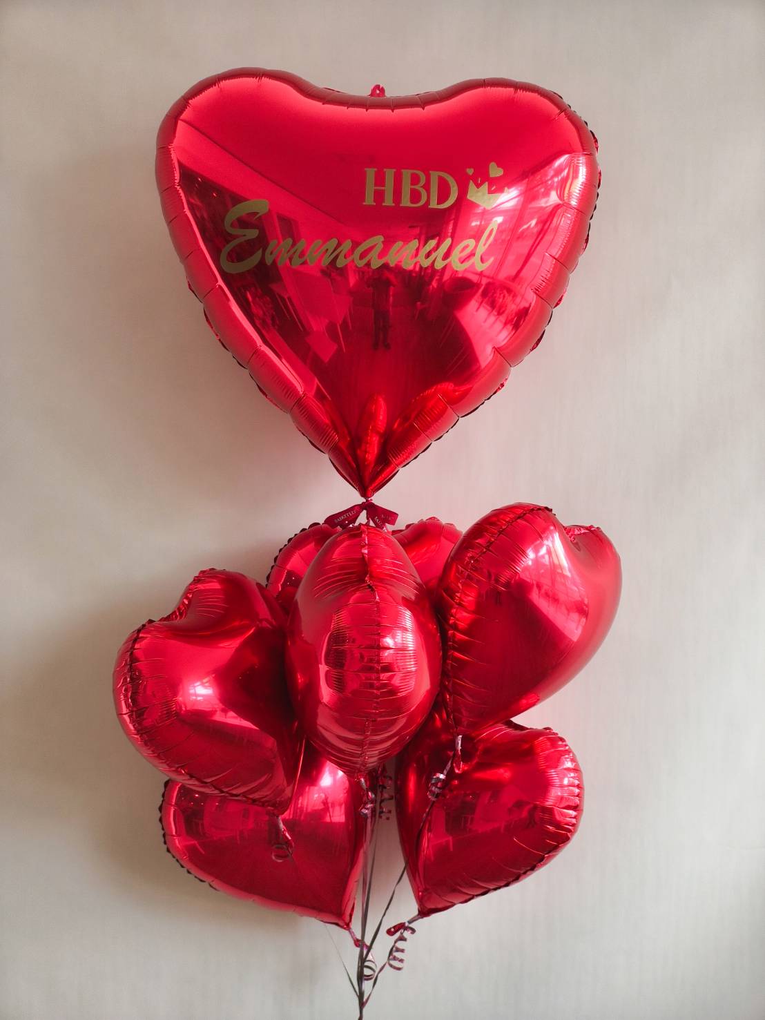 Bouquet of red heart-shaped foil balloons with a large personalised balloon reading “HBD Emmanuel” in gold letters.