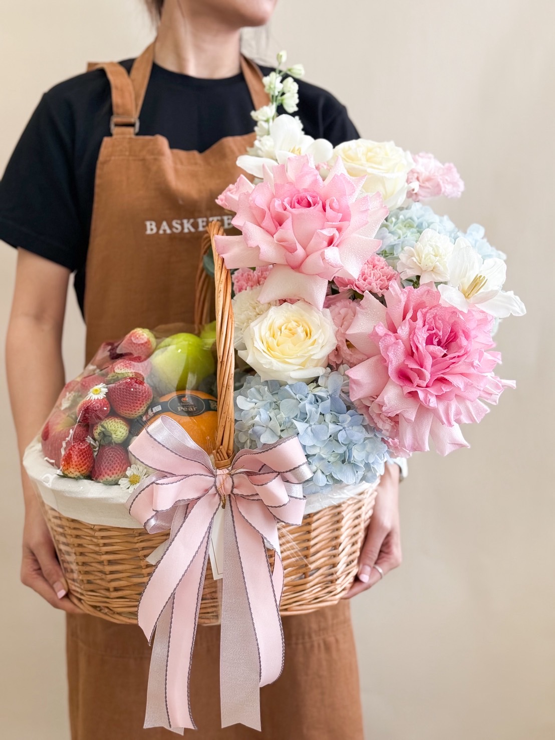 Wicker basket filled with pastel flowers including peonies, hydrangeas, roses, and fresh fruits like strawberries, pears, and oranges, tied with a pink ribbon.