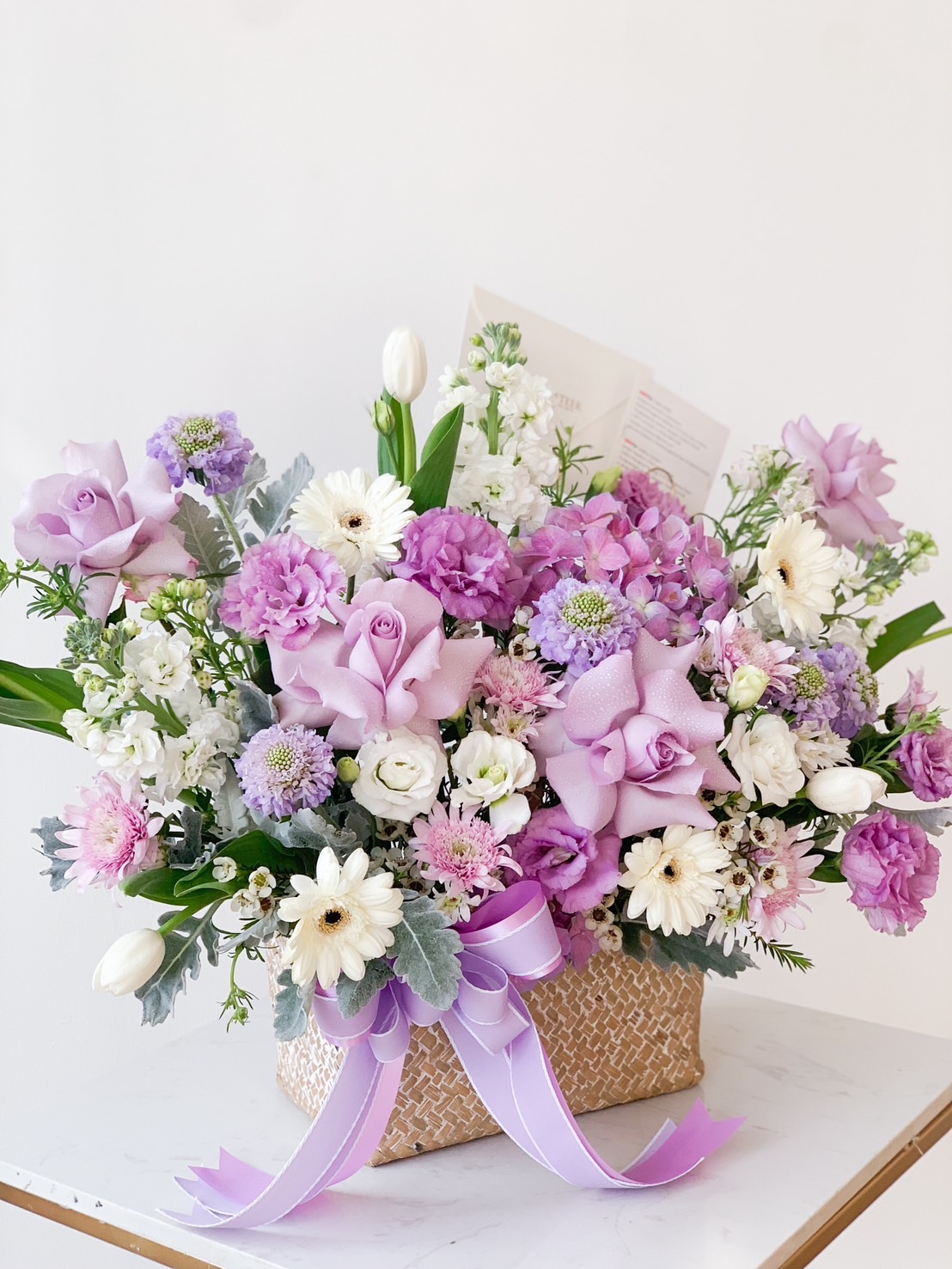 A woven basket filled with an elegant floral arrangement featuring purple roses, lavender carnations, white daisies, and green foliage. A lavender ribbon adorns the basket, adding a touch of charm. The background is a plain, off-white wall.