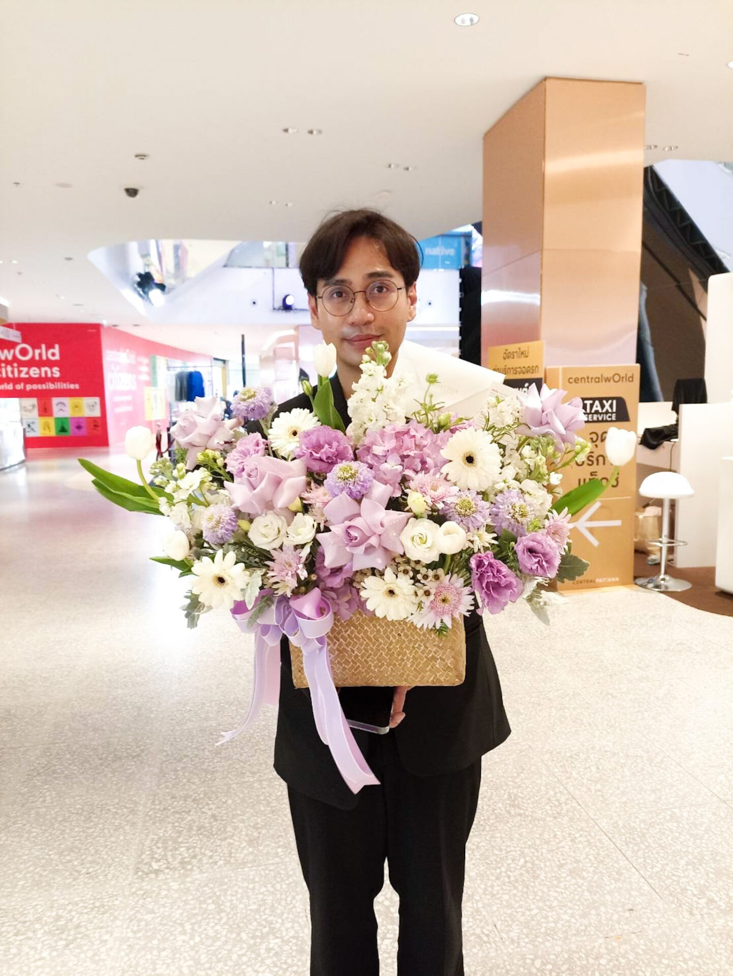 A person with short dark hair and glasses stands indoors, smiling while holding a large wicker basket filled with a colorful bouquet of flowers, including white and purple blooms. They wear a white shirt and black jacket, with a shopping mall background behind them.