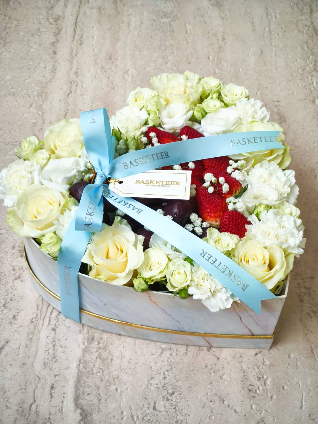Marble-patterned heart-shaped box filled with white roses, carnations, fresh strawberries, and blue Basketeer ribbon.