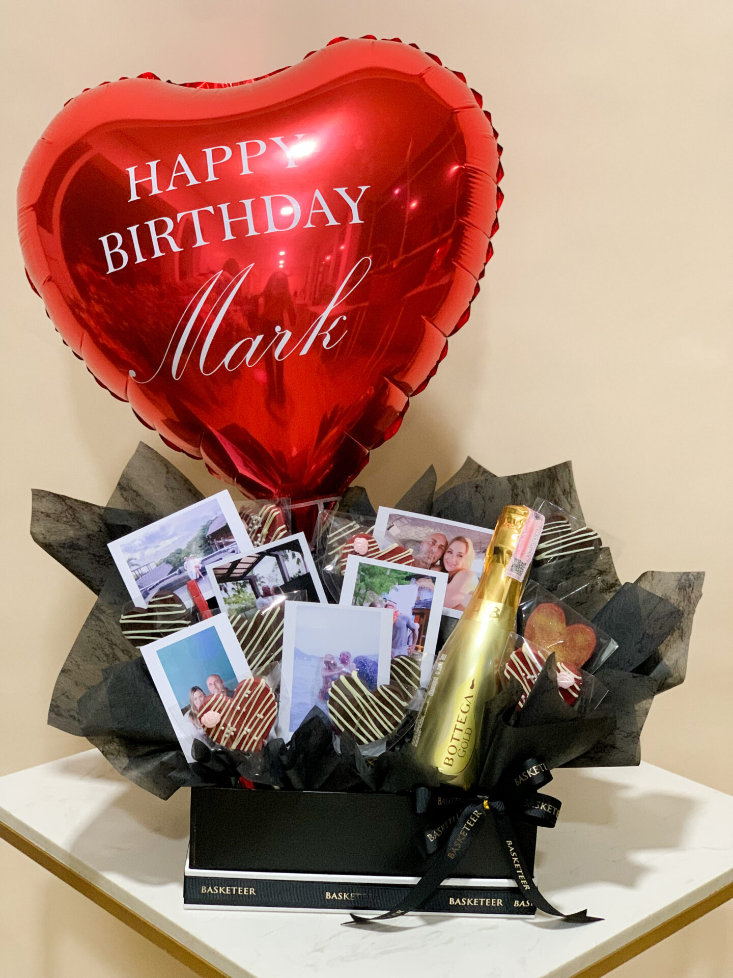 A luxurious heart-shaped red balloon, exquisitely adorned with the words "HAPPY BIRTHDAY Drawny" elegantly printed in white. The balloon features a sophisticated red ribbon and tassel gracefully cascading from the bottom, adding a touch of opulence. The scene is set against a pristine white paneled wall, further enhancing its charm. In the bottom left corner, the esteemed "Basketeer" brand logo proudly signifies quality and refinement.