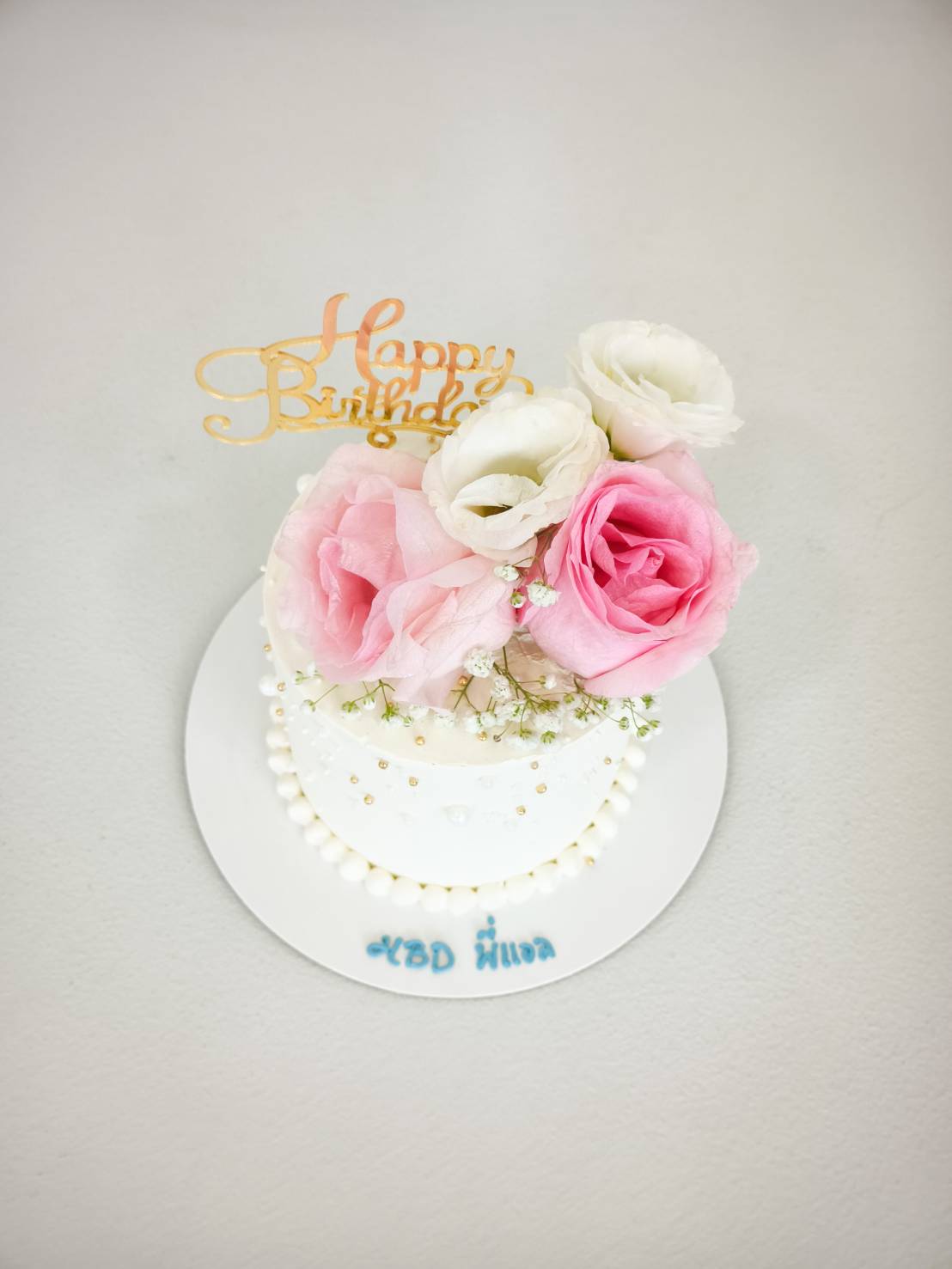 Elegant white birthday cake decorated with pink and white fresh roses, lisianthus, pearls, and a golden 