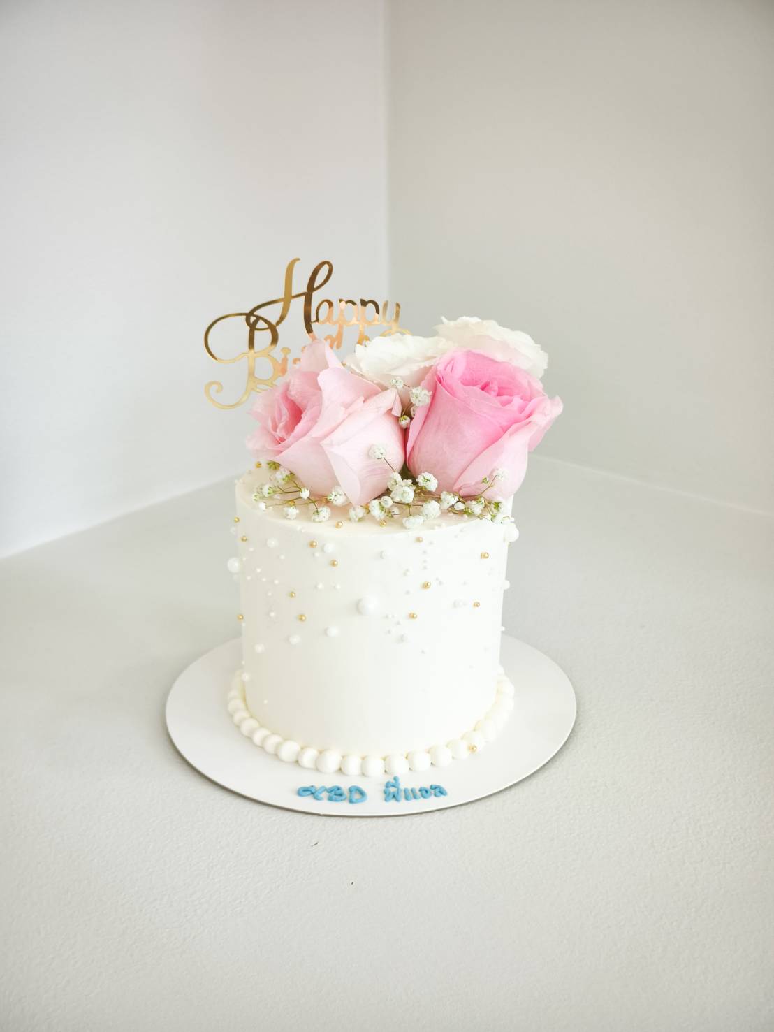 Elegant tall white birthday cake topped with fresh pink and white roses, baby’s breath, pearl decorations, and a golden topper.