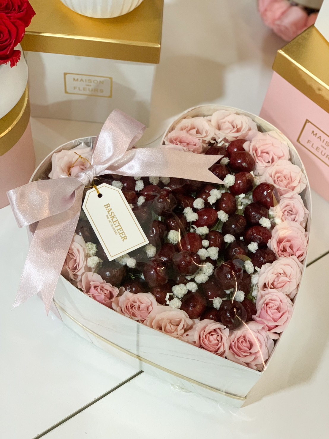 A heart-shaped gift box filled with fresh cherries, pink roses, and baby’s breath, wrapped with a pink satin ribbon and a gold-trimmed box.