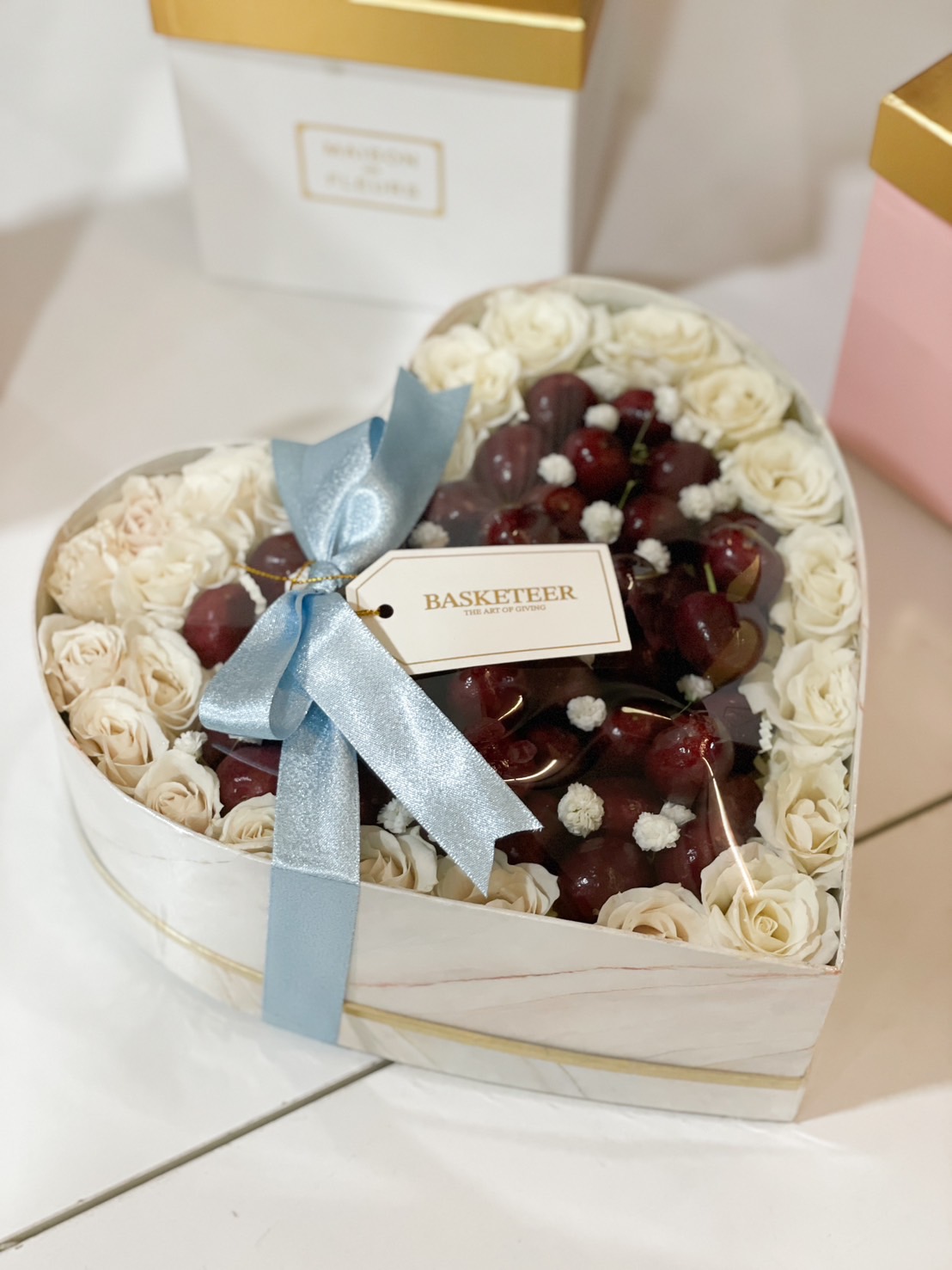 Heart-shaped gift box filled with white preserved roses and fresh cherries, tied with a blue satin ribbon and a Basketeer tag.