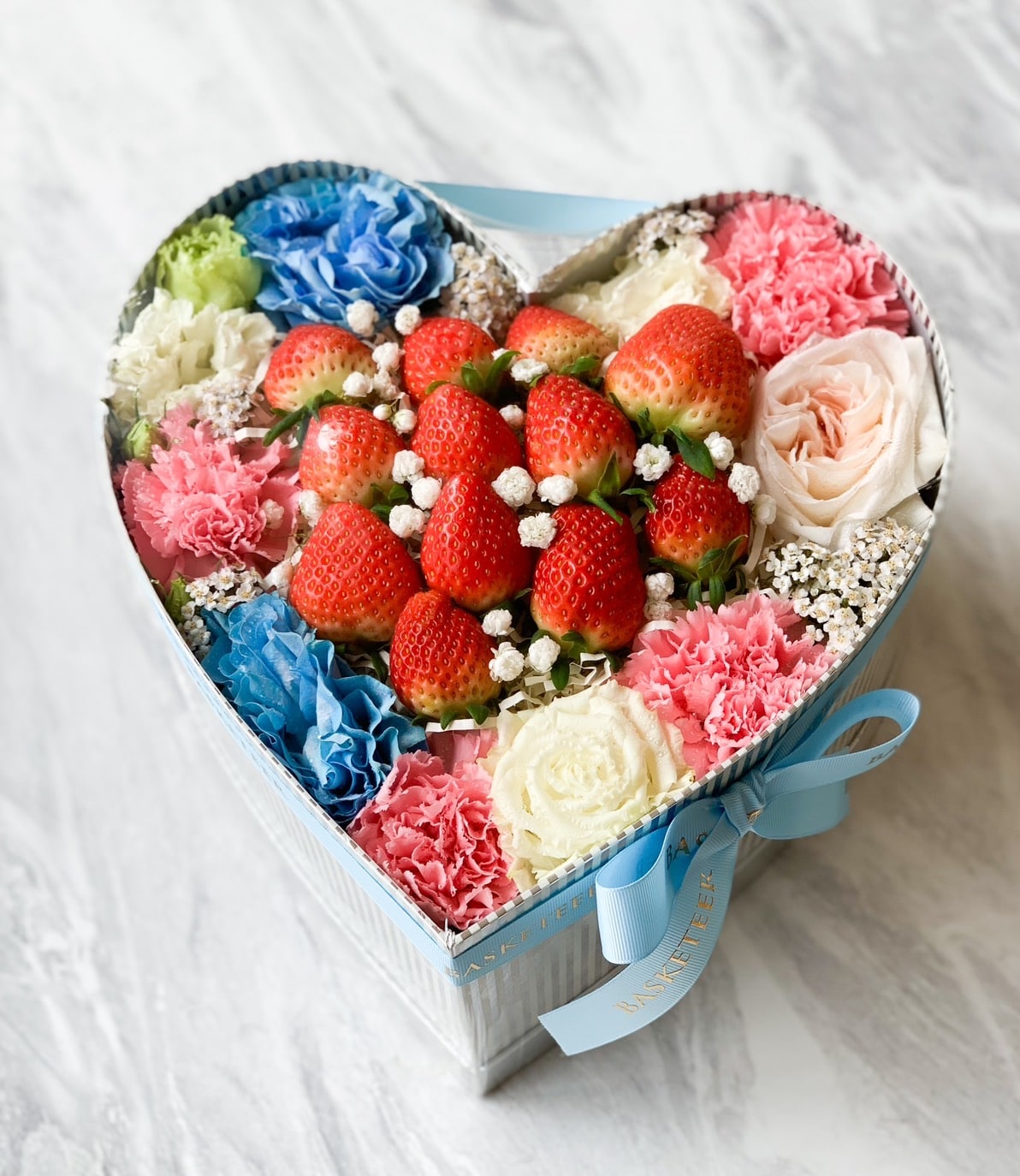 The Strawberry Romance Gift Box features a heart-shaped container with a pastel light blue exterior, filled with fresh strawberries at the center. It's surrounded by colorful flowers including pink, blue, white, and green carnations, as well as roses and baby's breath.