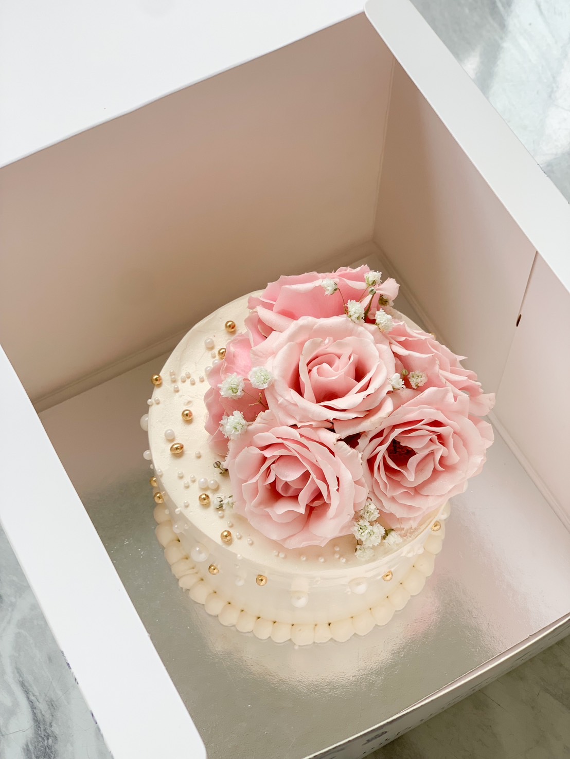 A sumptuous mini cake with delicate sweet roses, elegantly adorned with pink blossoms and petite white flowers, is showcased gracefully within a pristine white bakery box. This exquisite creation from Basketeer also features intricate gold and pearl-like embellishments along its edges. The decadent scene is set against a luxurious marble-like surface, evoking an air of timeless sophistication. A ribbon bearing the Basketeer logo adds a touch of exclusivity to this indulgent offering.