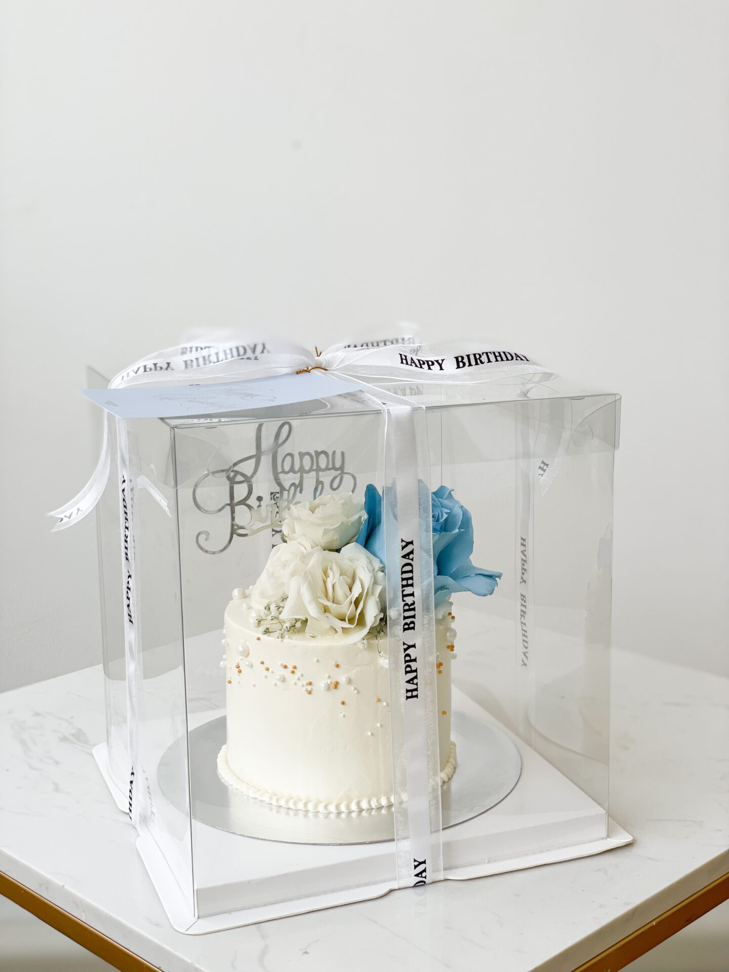 A decadent mini cake, elegantly adorned with intricate sweet roses and delicate white and blue flowers, is beautifully presented within a transparent, luxurious gift box. The box is meticulously tied with opulent ribbons inscribed with 