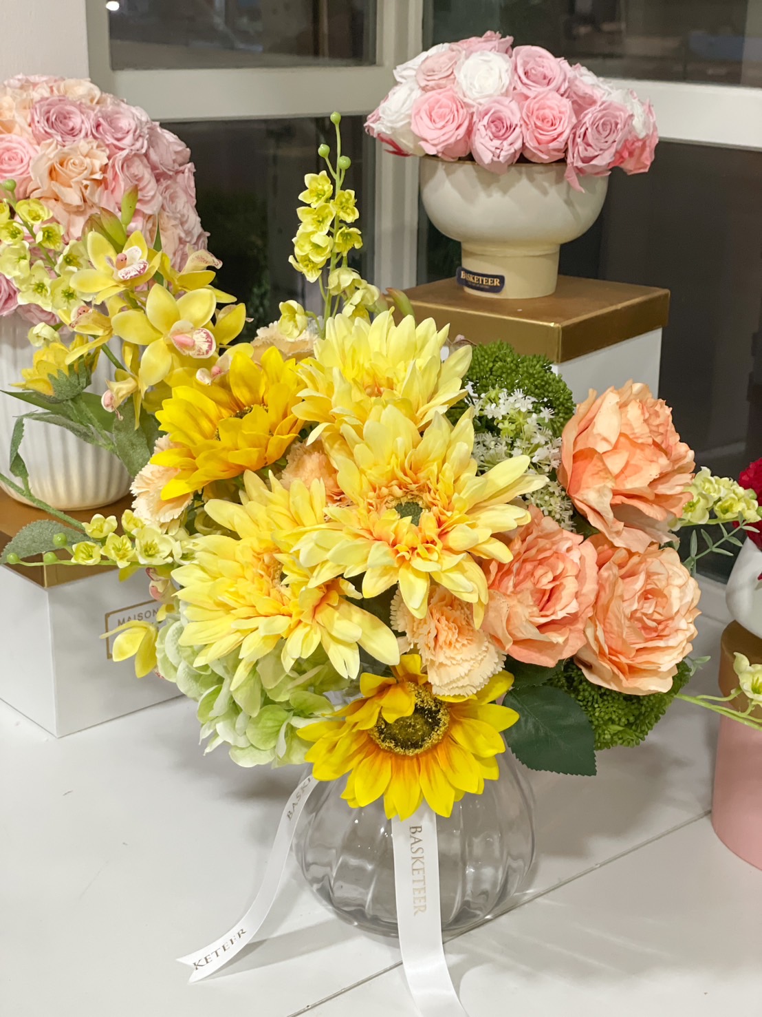 A beautiful artificial floral arrangement with sunflowers, yellow daisies, and peach roses in a chic glass vase, adding elegance to any space—ideal for home styling and gifting.