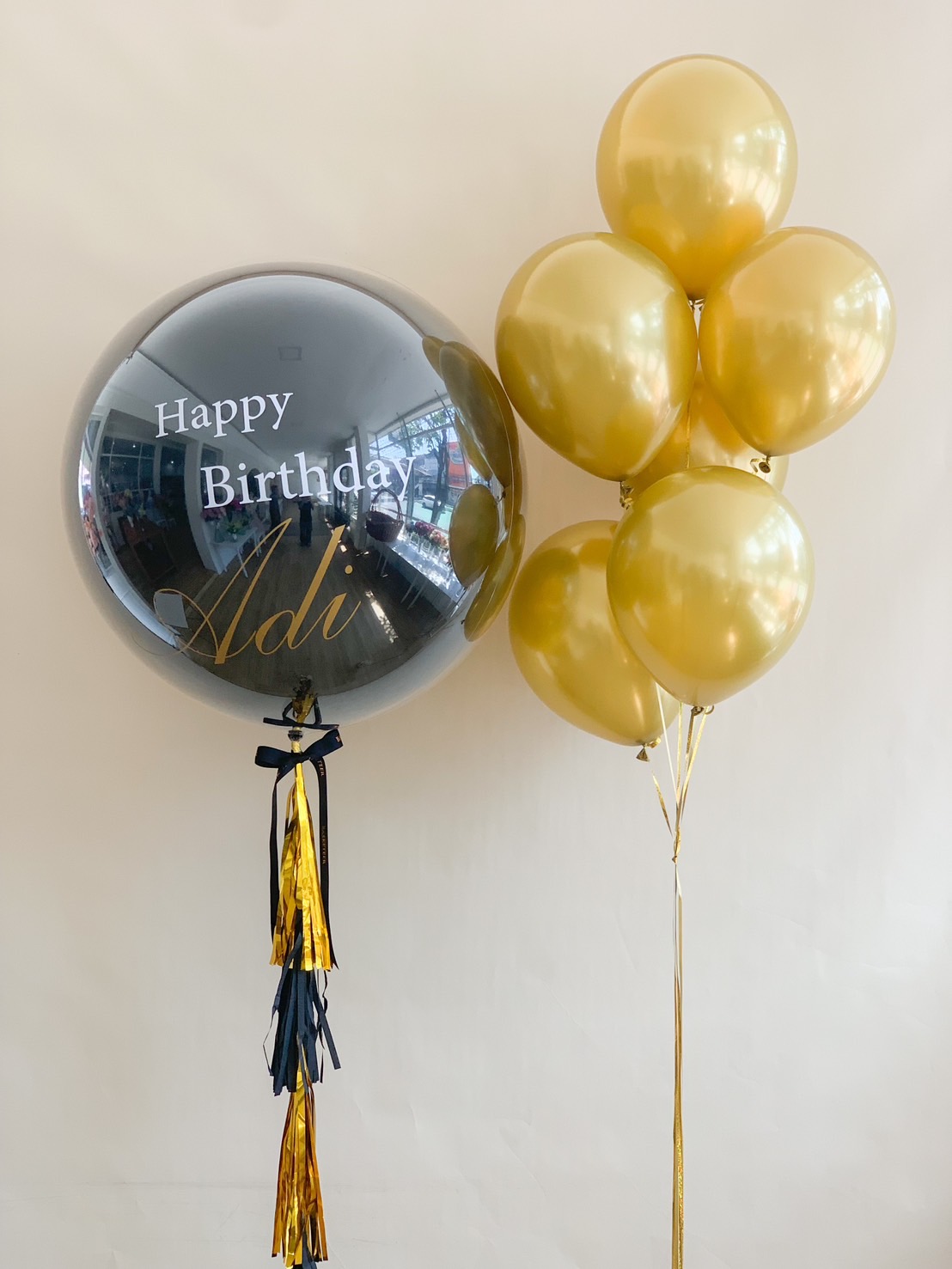 A premium black and gold birthday balloon set, featuring a large personalized black helium balloon with gold and white lettering, adorned with a stylish tassel tail, and paired with six metallic gold balloons. Perfect for an elegant birthday celebration.