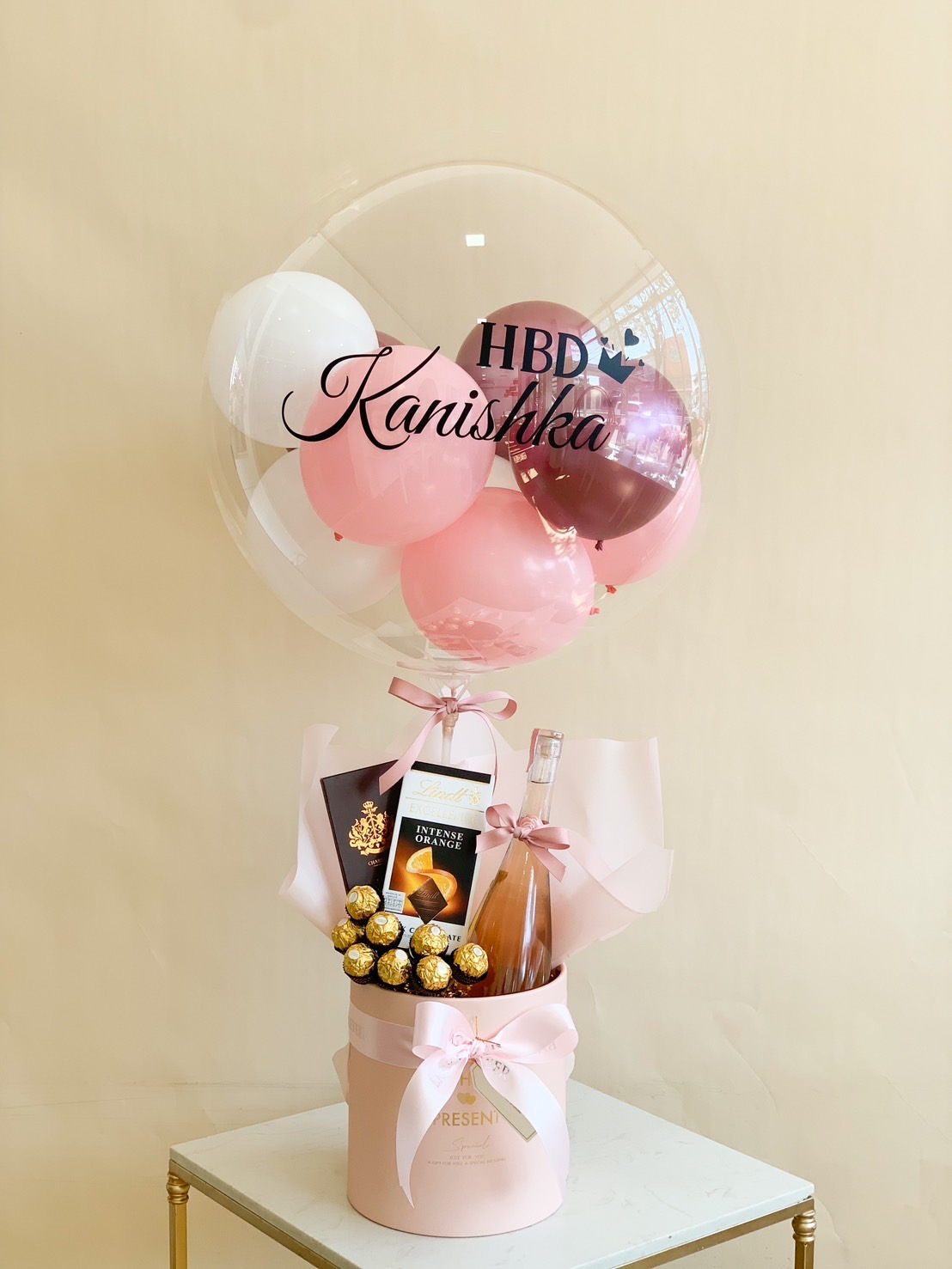 A stylish birthday gift set featuring a customized bubble balloon with pink, white, and metallic balloons inside, a premium bottle of rosé wine, Lindt intense dark chocolate, and Ferrero Rocher chocolates, elegantly arranged in a luxury round gift box with pink ribbon—perfect for birthday surprises and celebrations.