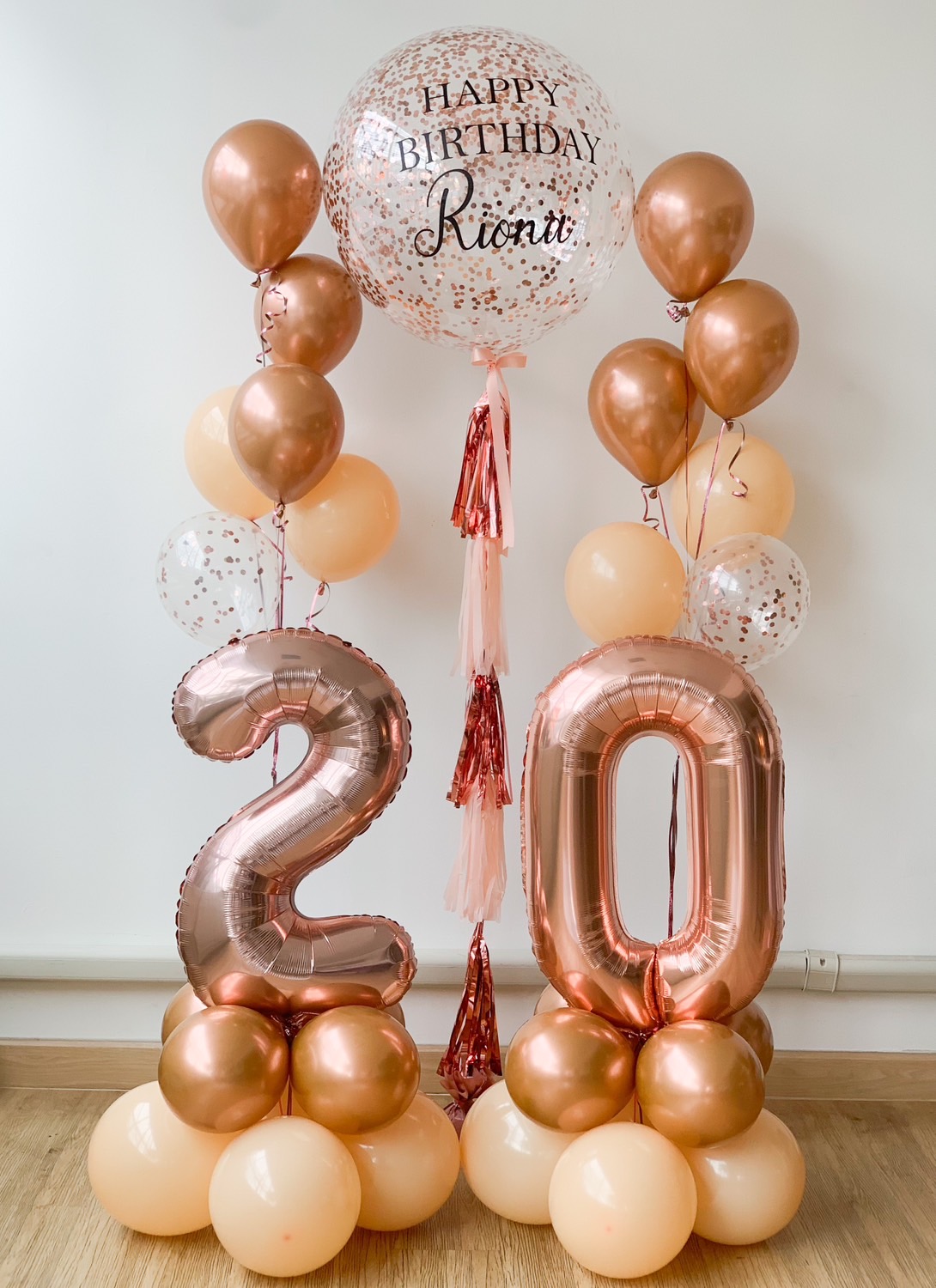 A festive balloon arrangement featuring large rose gold foil balloons forming the number 20, set on a base of smaller peach and rose gold balloons. Above them, a transparent balloon with confetti inside displays the text 