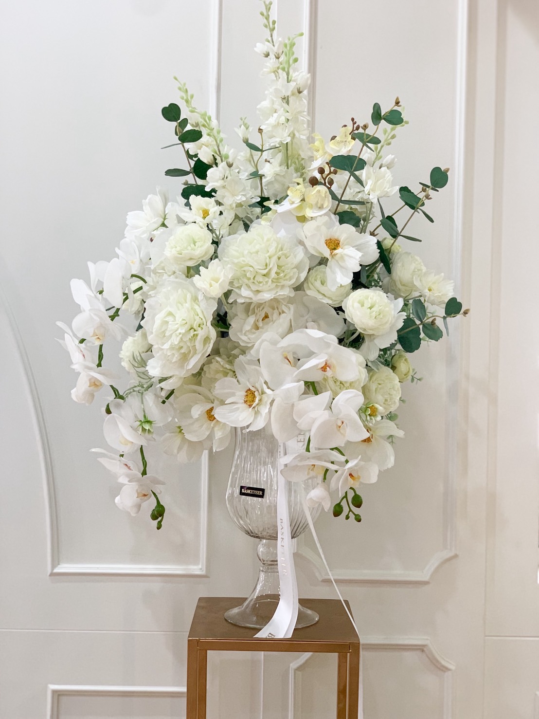 A luxury white flower vase arrangement with white orchids, peonies, delphinium, and greenery in a clear glass vase