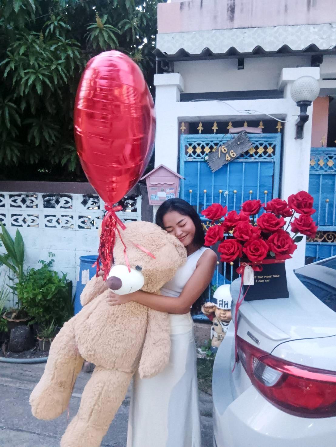 A woman stands outside next to a car, smiling and hugging a large teddy bear. She holds a red heart-shaped balloon. On the car's trunk is a bouquet of red roses. A house with a blue gate and lush greenery is in the background.