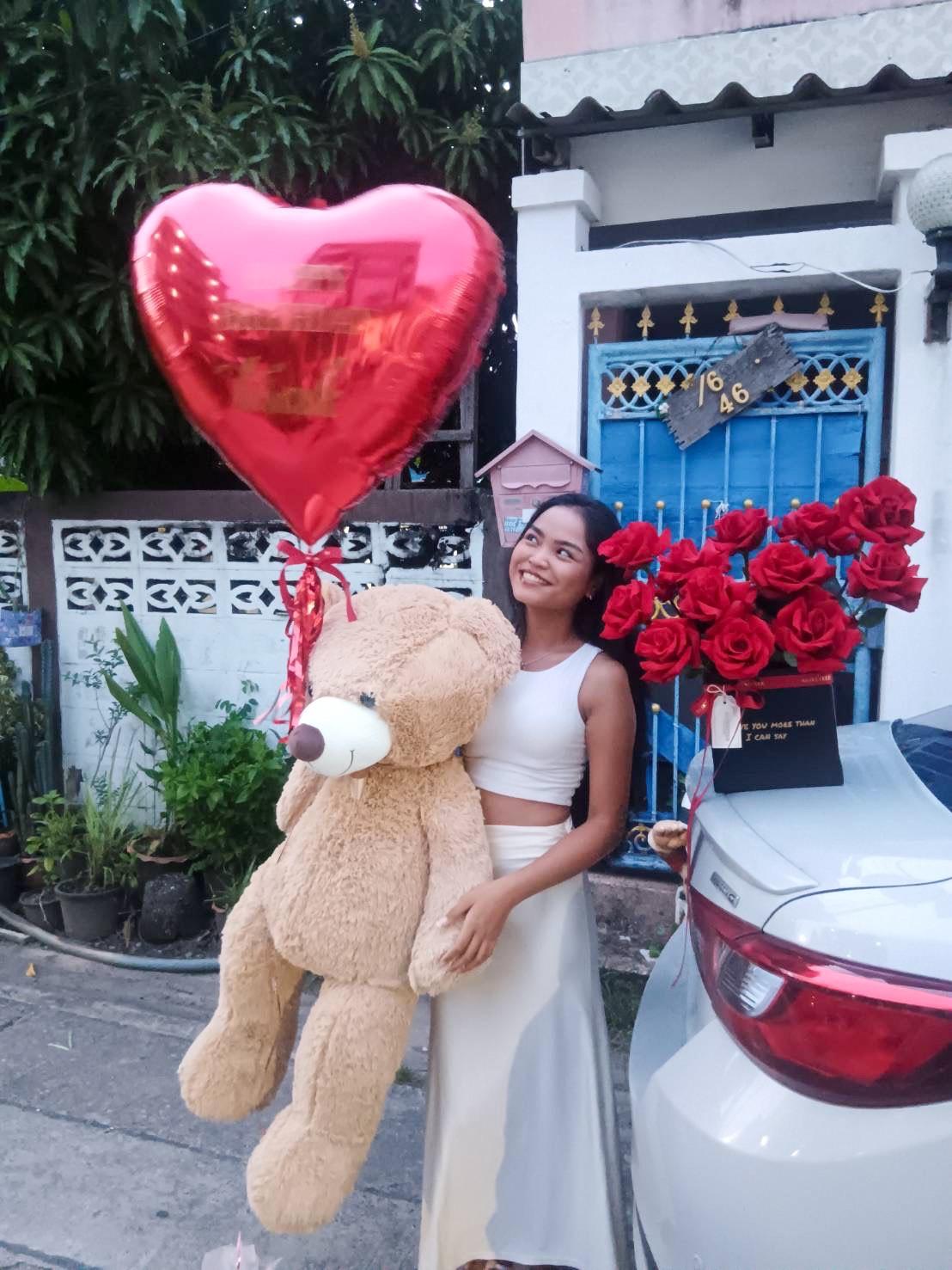 A woman in a white outfit stands outside next to a car holding a large teddy bear and a heart-shaped red balloon. A bouquet of red roses is placed on the car's trunk. The background features a house with a blue door and potted plants.