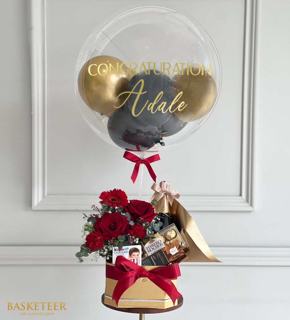 A luxurious congratulatory gift arrangement showcases a pristine clear balloon filled with elegant gold and black balloons, beautifully adorned with the personalized message 