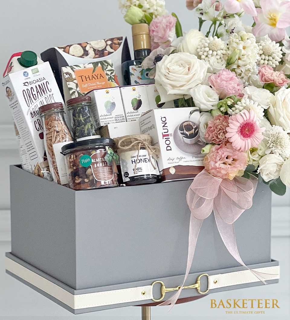 A sumptuous gift basket curated by Basketeer, featuring an array of exquisite organic products. Included are a premium carton of coconut milk, a jar of golden honey, a bottle of the finest olive oil, select gourmet nuts and rich chocolates, all complemented by an assortment of specialty teas and aromatic coffee. The basket is elegantly adorned with a pink ribbon and a floral arrangement of delicate white and blush blooms. The prestigious Basketeer logo is proudly displayed, symbolizing luxury and thoughtful gifting.