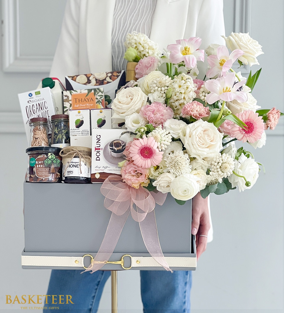 A person gracefully holds a sumptuous Basketeer gift basket overflowing with an exquisite selection of gourmet delicacies and an enchanting array of pastel-hued fresh flowers. The basket is elegantly adorned with a soft pink bow and features premium items such as artisanal cookies, golden honey, and fine tea, all meticulously curated for an indulgent experience. The distinguished 