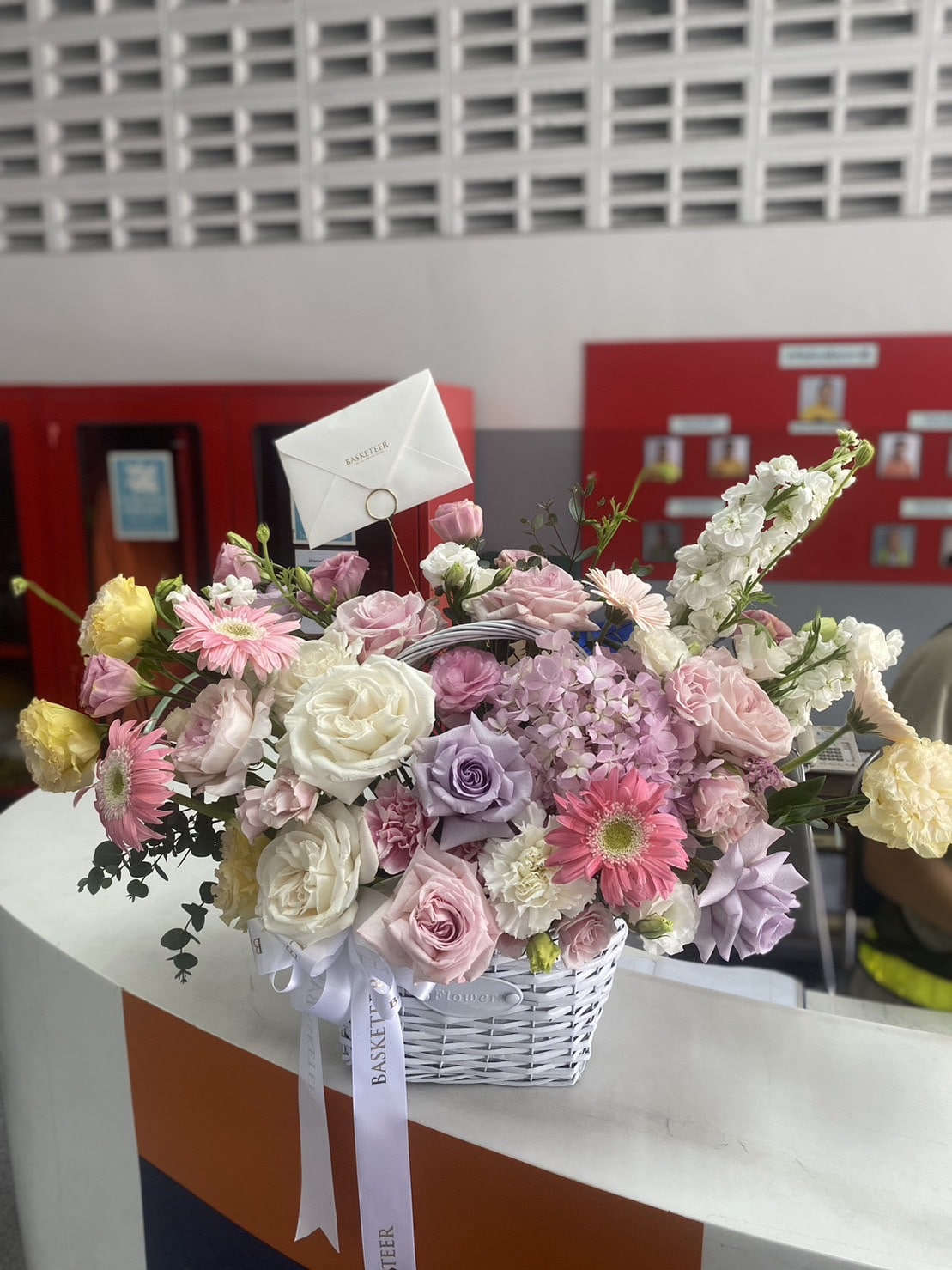 A white wicker basket filled with a vibrant assortment of flowers including pink, yellow, and purple roses, gerbera daisies, and white lilies. A small white envelope is nestled among the flowers, and the basket is adorned with a white 