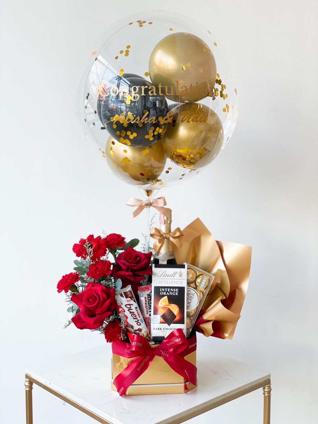 A celebration gift set featuring a transparent balloon with gold and black smaller balloons inside, bearing the words "Congratulations." The set includes red roses, a bottle of wine, and various chocolates, all elegantly arranged with gold wrapping and red ribbons.