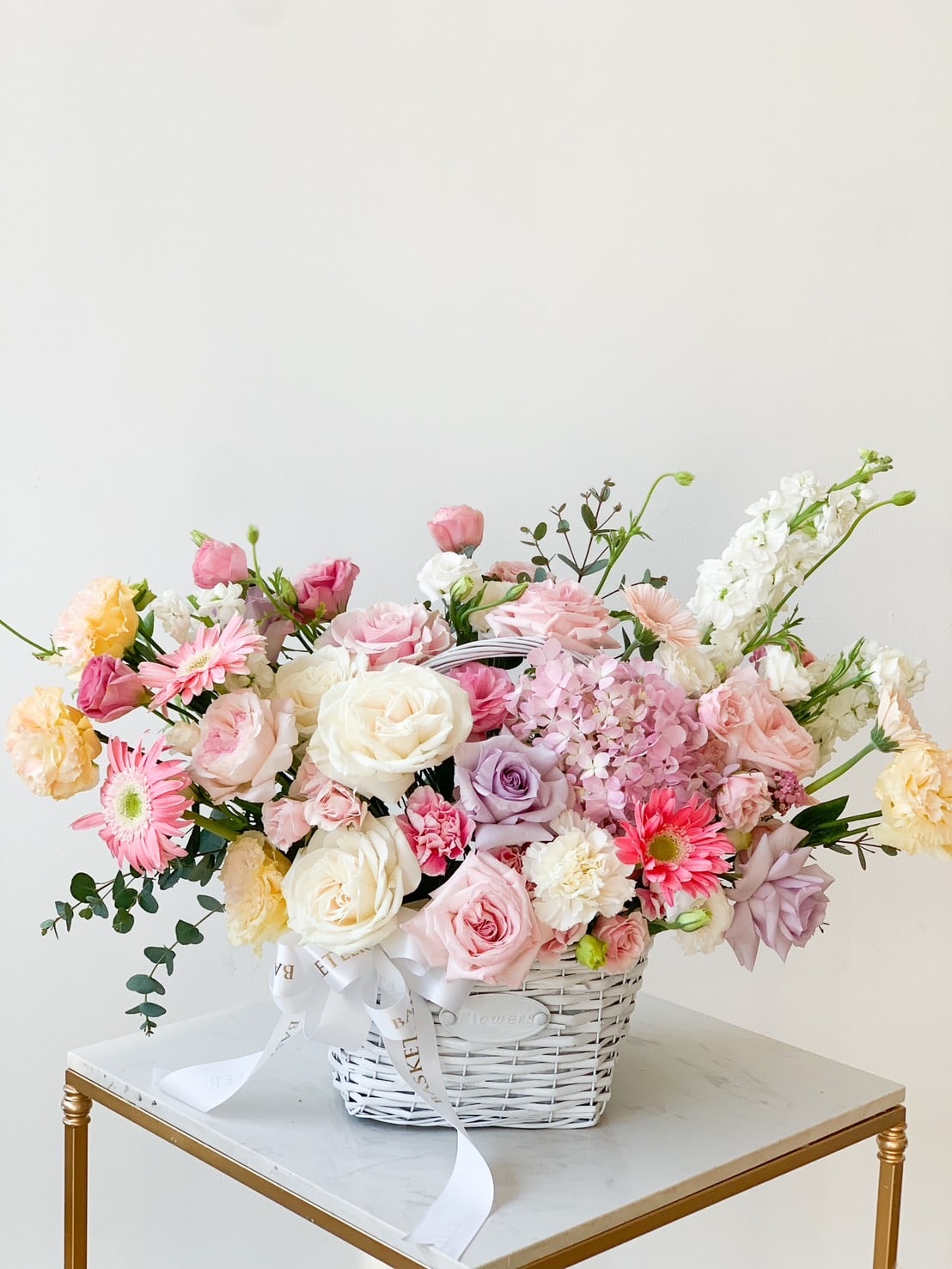 A white wicker basket filled with an assortment of flowers, including pink roses, white roses, light pink gerberas, and purple hydrangeas, placed on a small marble-top table with a golden frame. A white ribbon is tied around the basket handle.