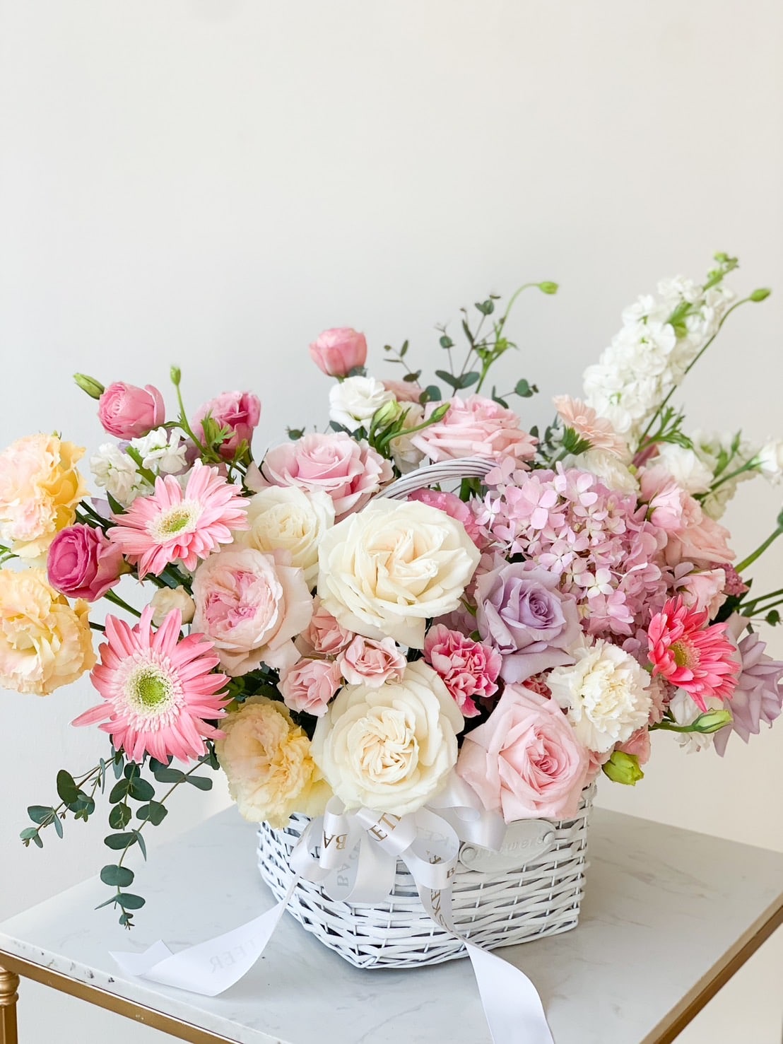 A white wicker basket filled with a beautiful arrangement of various flowers, including pink gerbera daisies, white roses, and purple hydrangeas, set on a white tabletop against a plain white background.