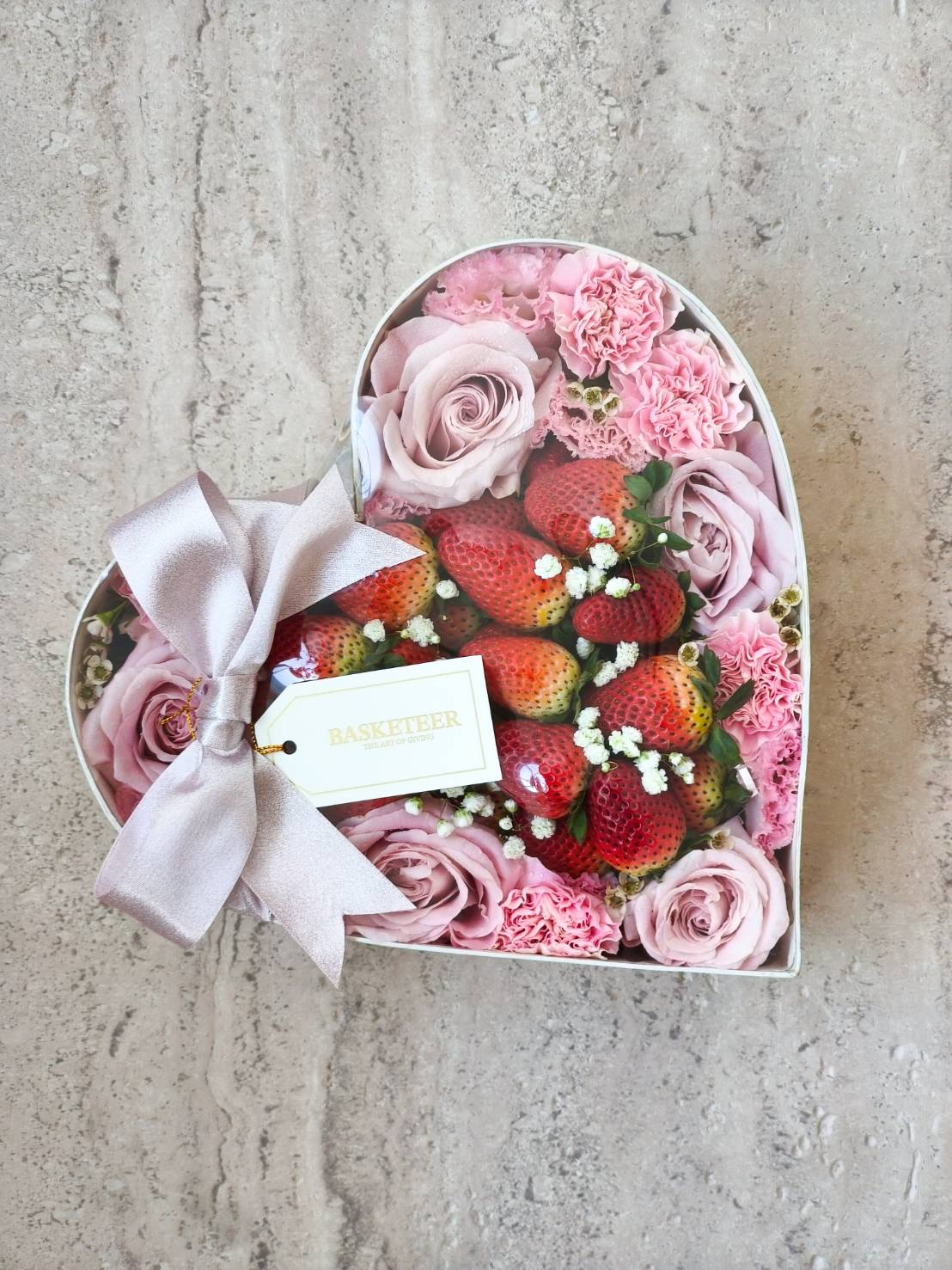 Heart-shaped gift box with pink roses, carnations, and fresh strawberries, decorated with satin ribbon and Basketeer card.