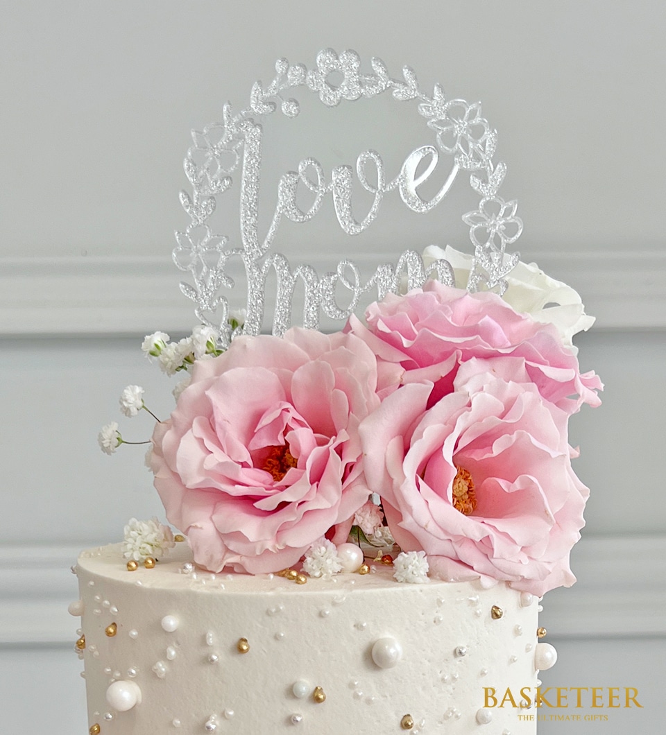 A luxurious white mini cake, elegantly adorned with large pink roses and delicate white flowers. Pearl-like beads embellish the cake, adding a touch of sophistication. Crowned with a floral wreath ornament that beautifully spells 